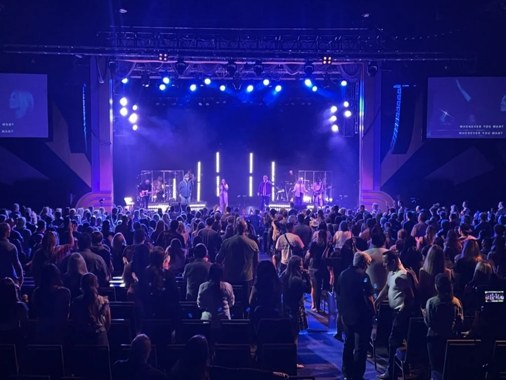 Close-up shot of a professional video camera capturing a colorful stage performance with pink, purple, and blue lighting, with the camera's screen showing the scene and the subtitle 'Mucha iluminación'.
