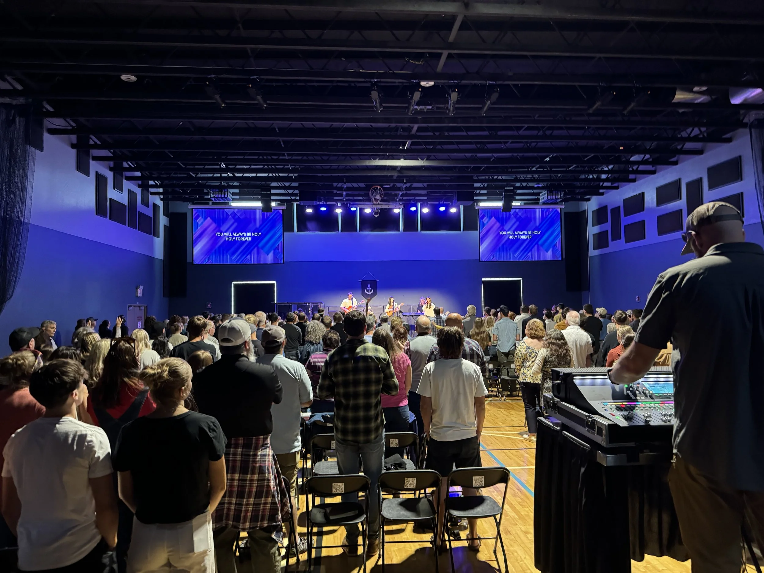 An empty indoor auditorium with a stage, a large projection screen, and rows of chairs facing the stage. The wall has a series of acoustic panels arranged in a grid pattern.