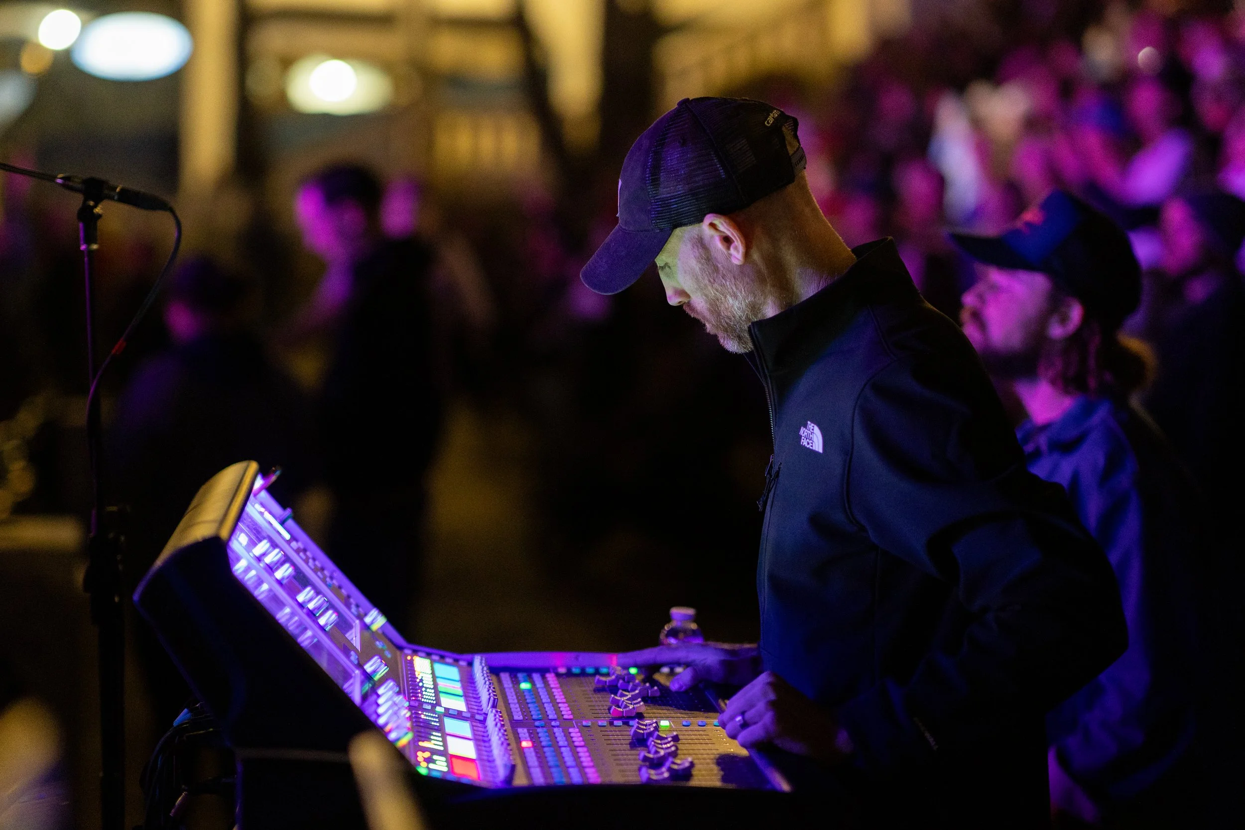 A sound engineer operates a digital mixing console with colorful lights at a concert, while the stage in the background is illuminated and a crowd of people watches.