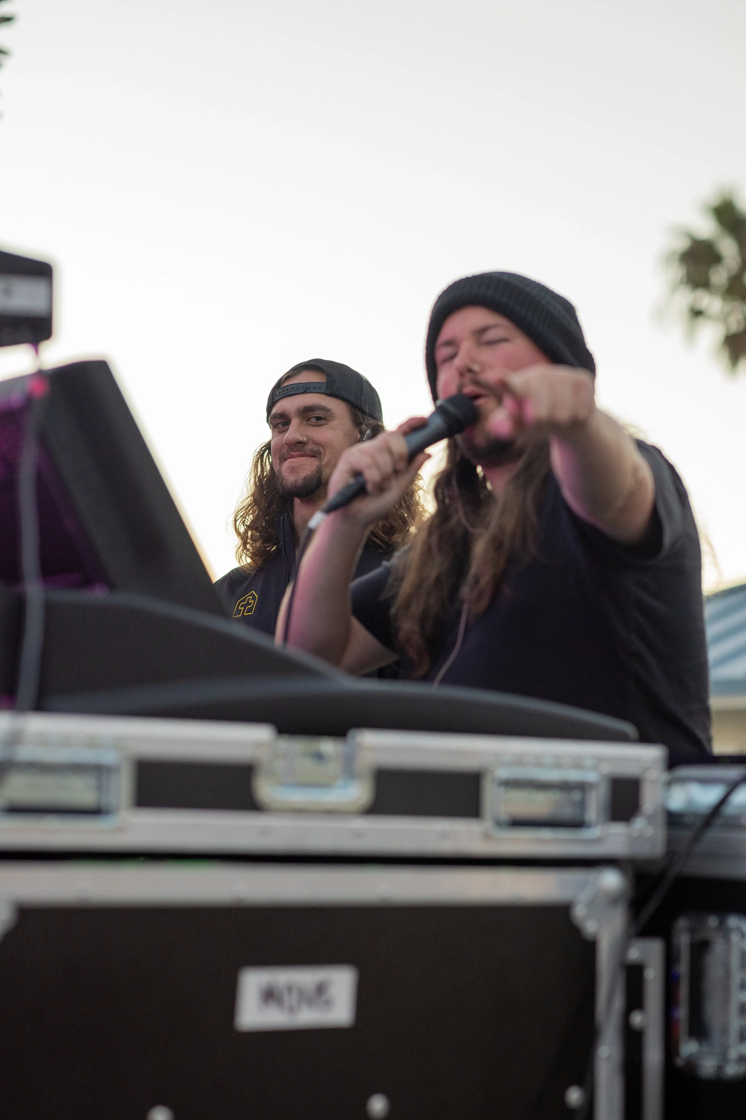 Two men, one with long hair and a beard, and the other with long hair and a beard, are performing as DJs outdoors, with one holding a microphone and the other smiling, against a clear sky and some trees in the background.