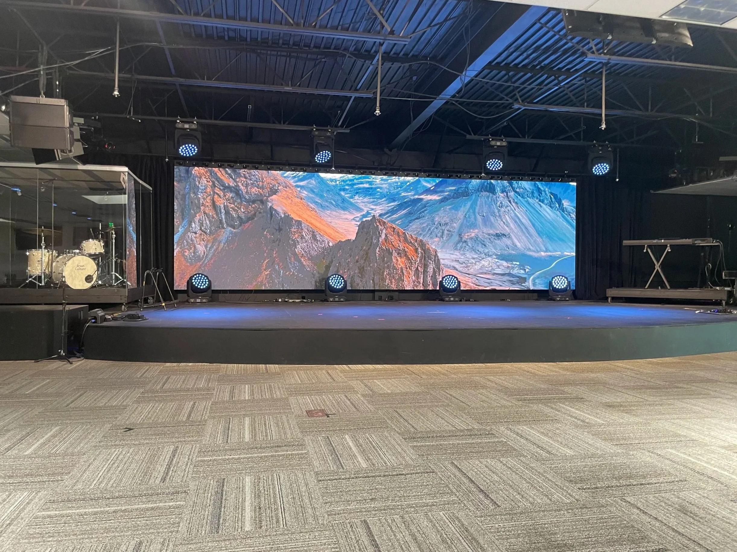 A band performing on stage with a large screen in the background displaying the word 'JOY' and a smiling face. The lead singer is holding a microphone, raising his hand, and singing passionately. Other band members are playing guitars and singing.