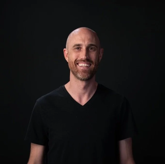 A smiling bald man with a beard wearing a black V-neck T-shirt against a dark background.