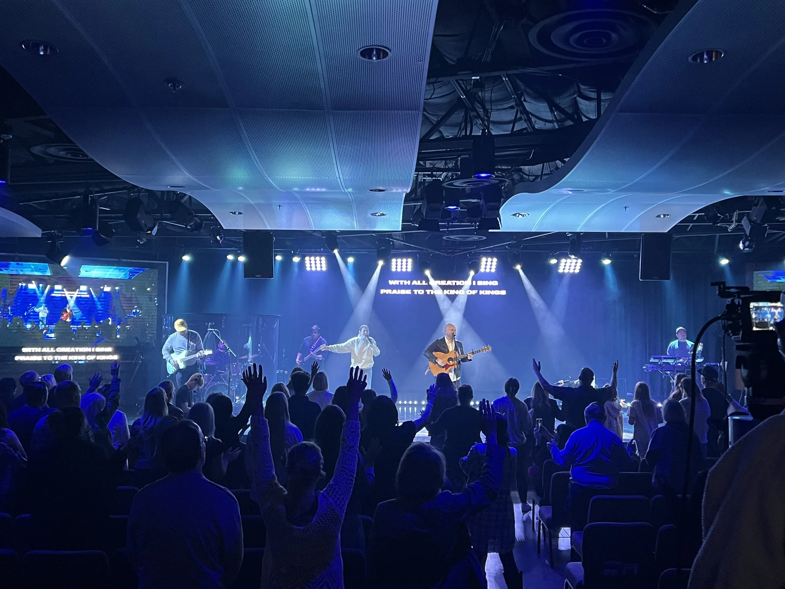 A live worship concert with a band performing on stage. The stage has multiple musicians, including a singer with a guitar, surrounded by colorful stage lights, and an audience with hands raised in front of the stage.
