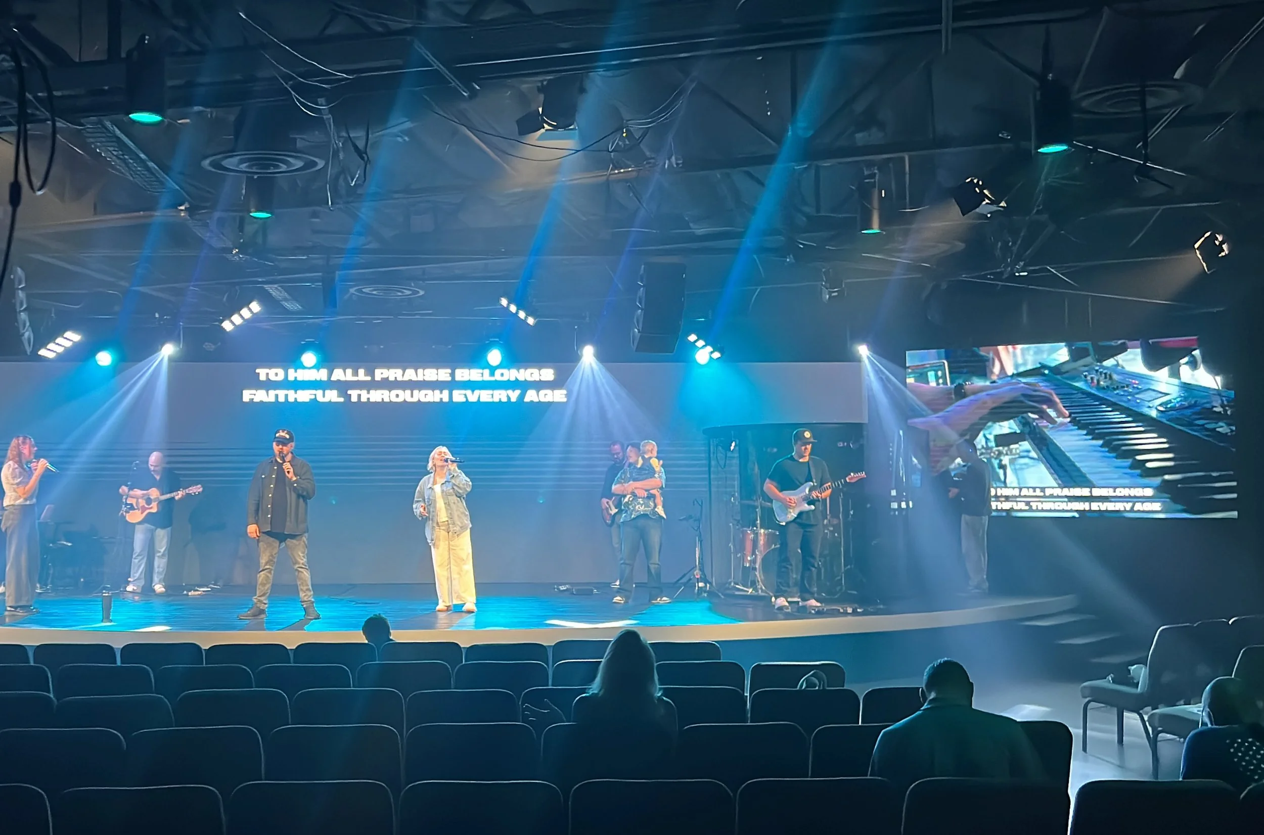 A live worship band performing on stage with blue spotlights, a large screen displaying lyrics, and a few audience members seated watching.