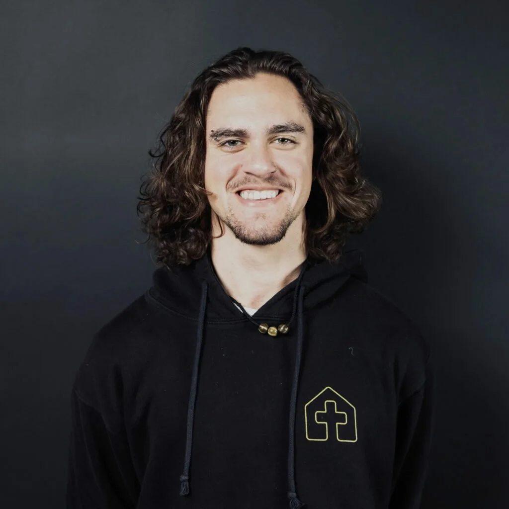 Young man with long, curly hair, smiling, wearing a black hoodie with a yellow logo on the chest, against a dark background.