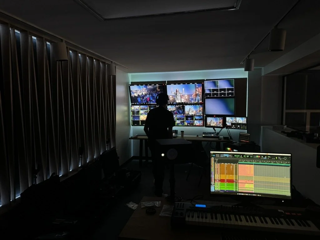 A man in a black shirt with a tattoo on his neck is working as a DJ in a studio, wearing a headset with a microphone. In the background, multiple screens display a woman with curly hair and glasses, appearing to be speaking or gesturing with her hands during a virtual event or recording.