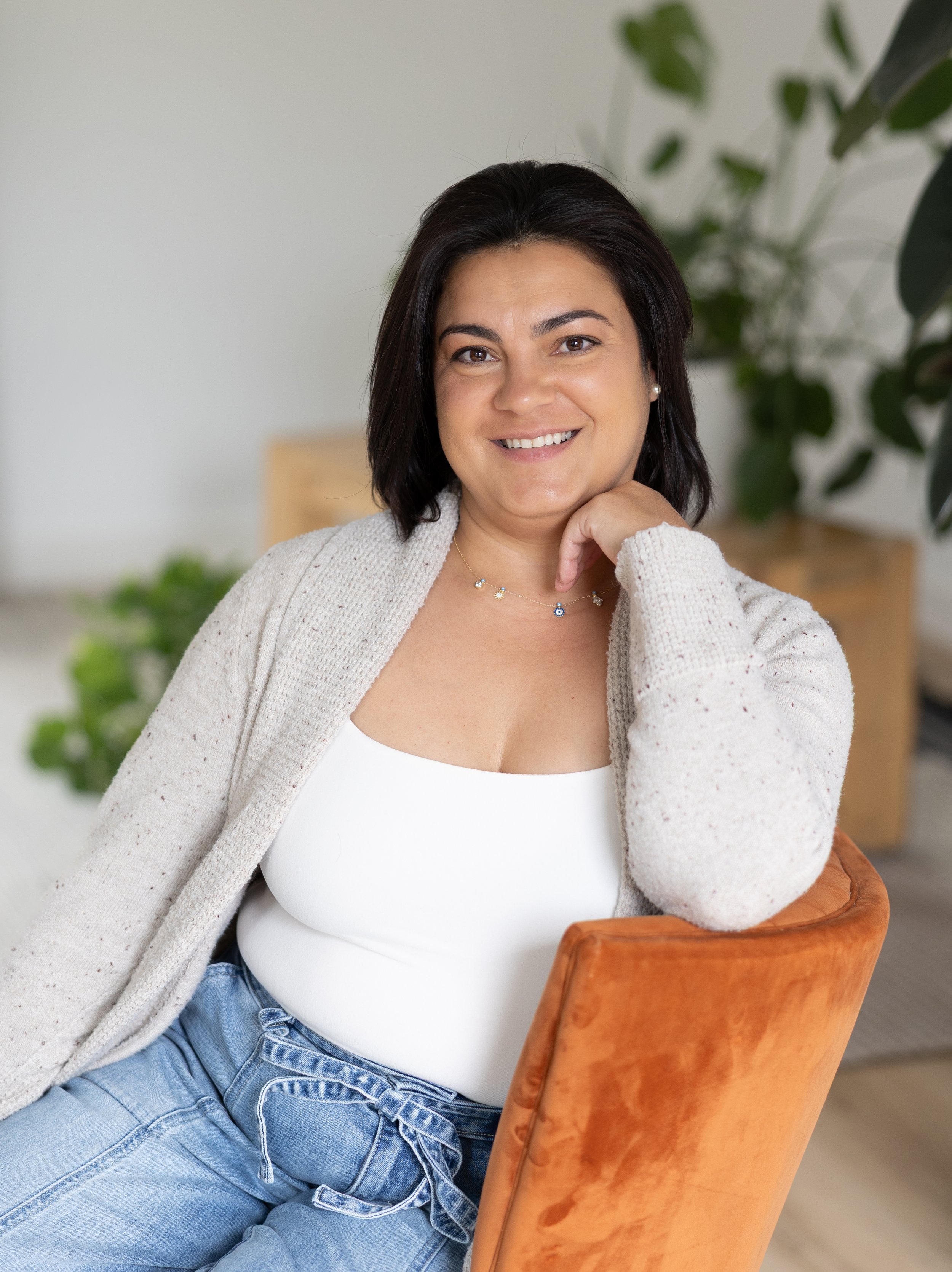A woman with dark hair and a white top sitting in a wooden chair, smiling, with a background of green plants and a light-colored room.