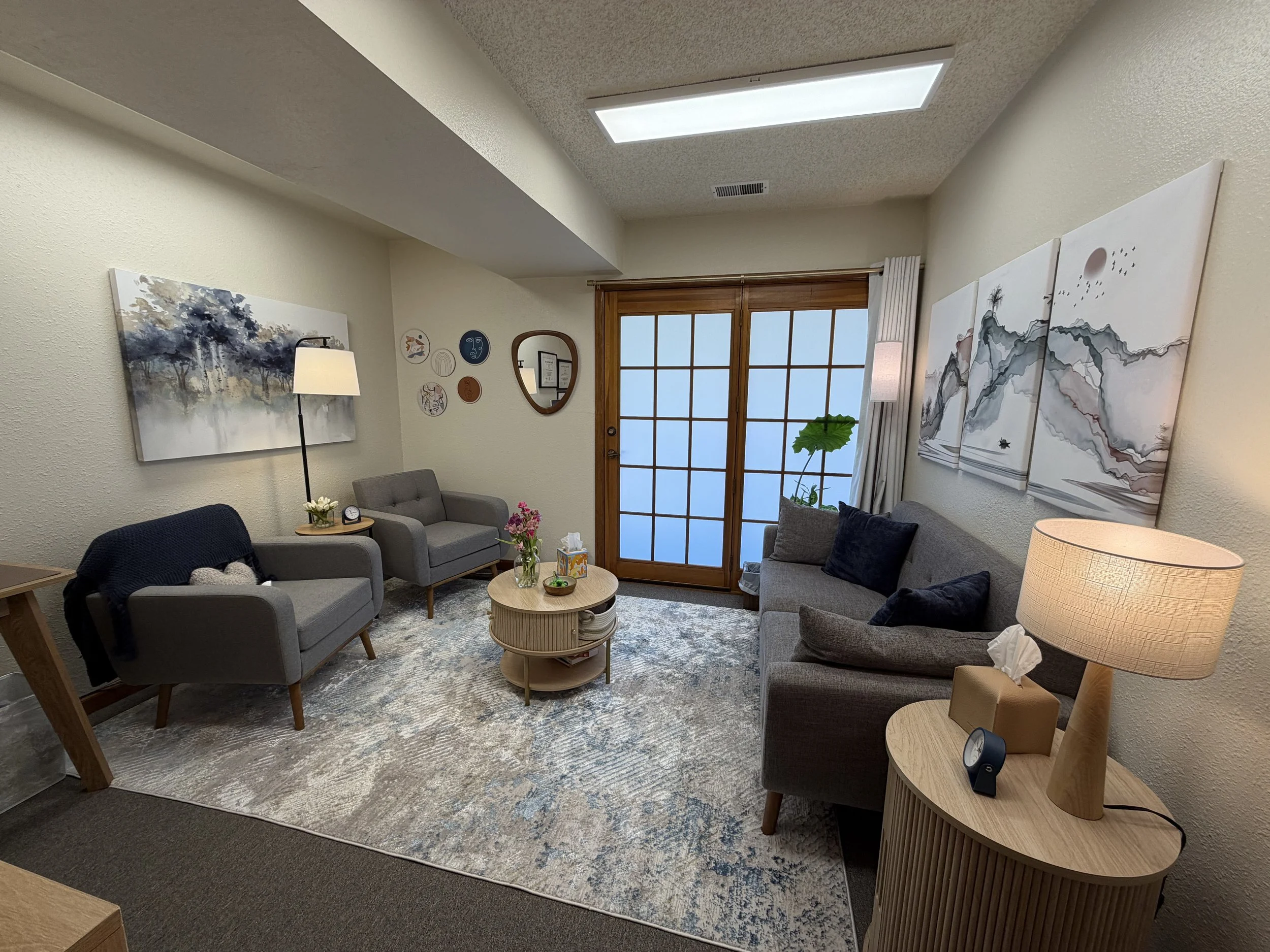 A cozy office with gray sofas, a wooden coffee table with flowers and tissues, a tall floor lamp, wall art, a sliding glass door with wooden frames, a potted plant, and a textured ceiling with a rectangular light fixture.