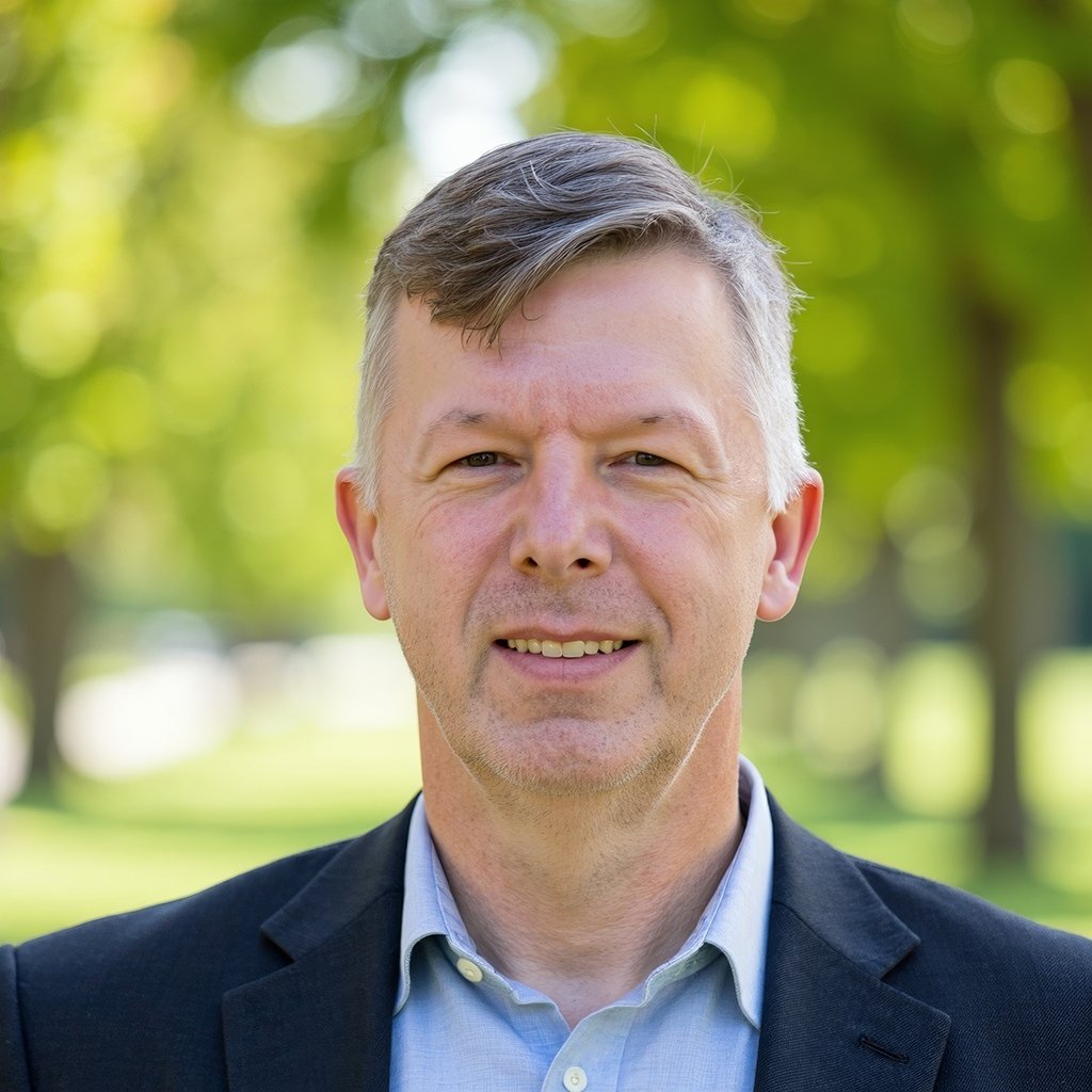 Portrait of a man with gray hair and blue eyes, smiling, wearing a blue collared shirt and a dark blazer, standing outdoors with green trees and blurred background.