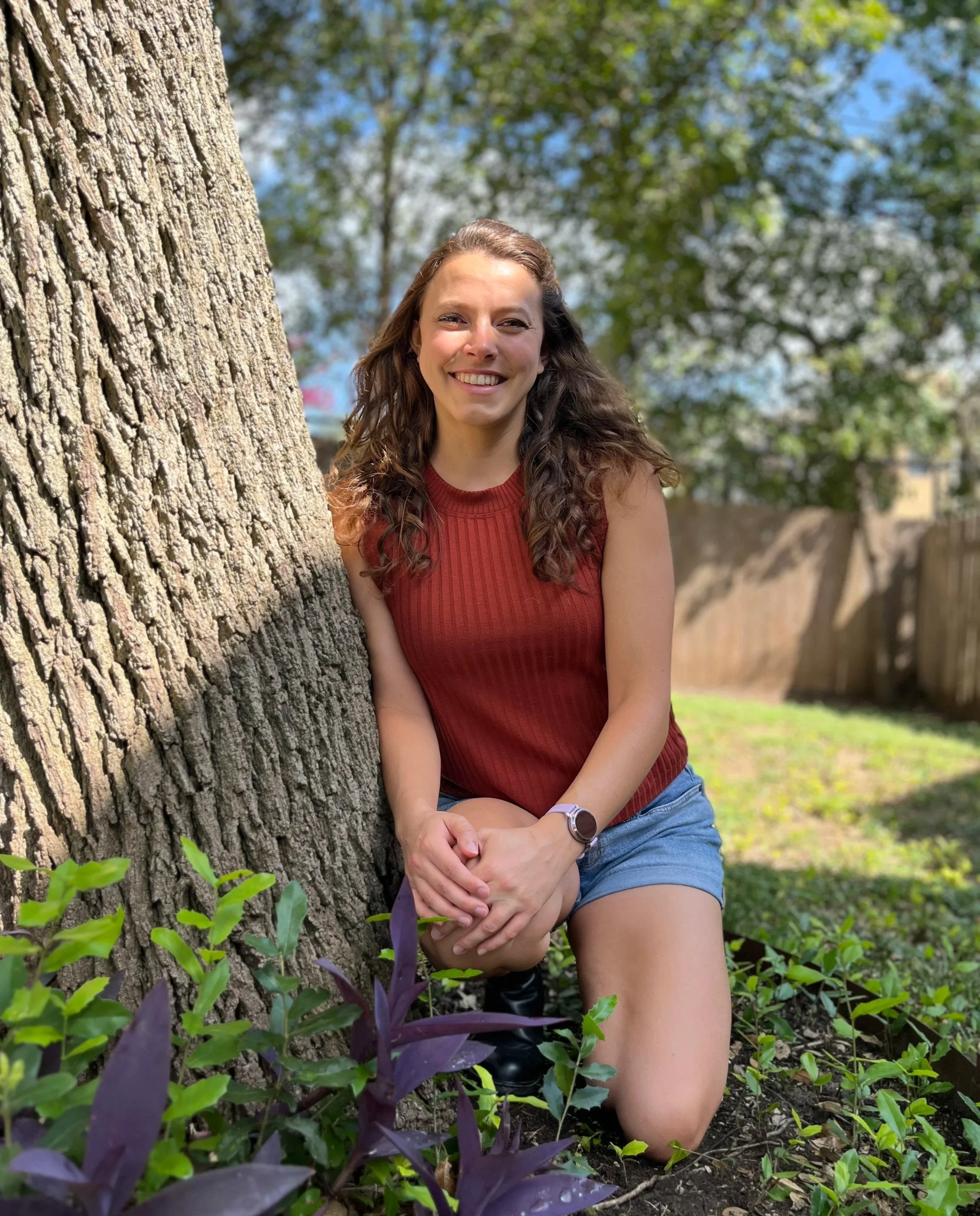 Mary Joncas in a kneeling pose next to a tree with small foliage around.