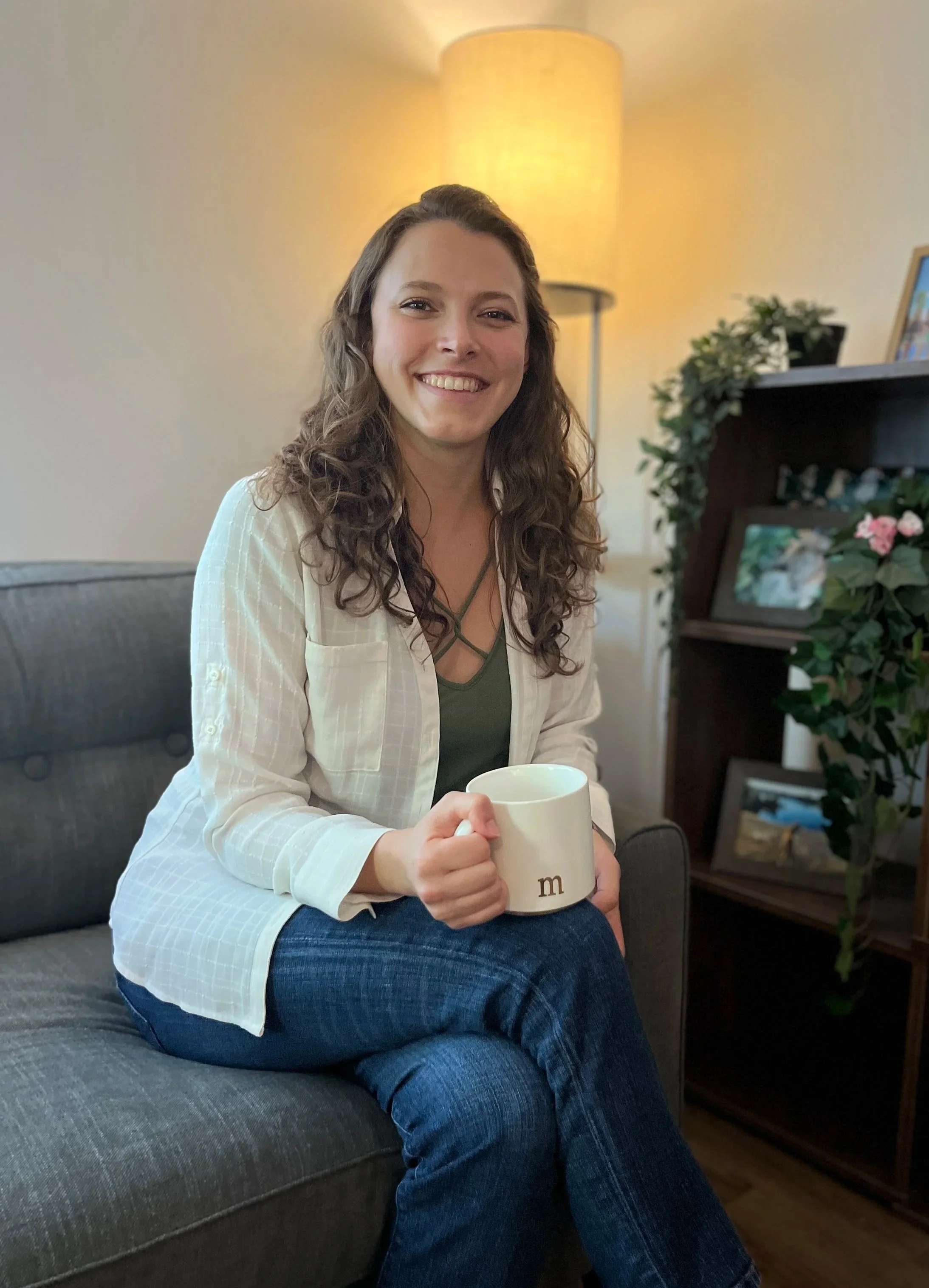 Mary Joncas smiling and sitting on a gray couch, holding a coffee mug. There is a standing lamp behind her and a wooden bookshelf with framed photos and plants to her right.