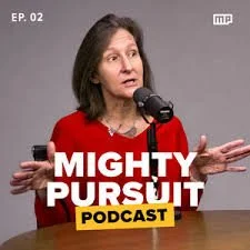 A woman with shoulder-length brown hair speaking into a microphone, wearing a red top, in front of a gray background. Text overlay says 'MIGHTY PURSUIT PODCAST'.