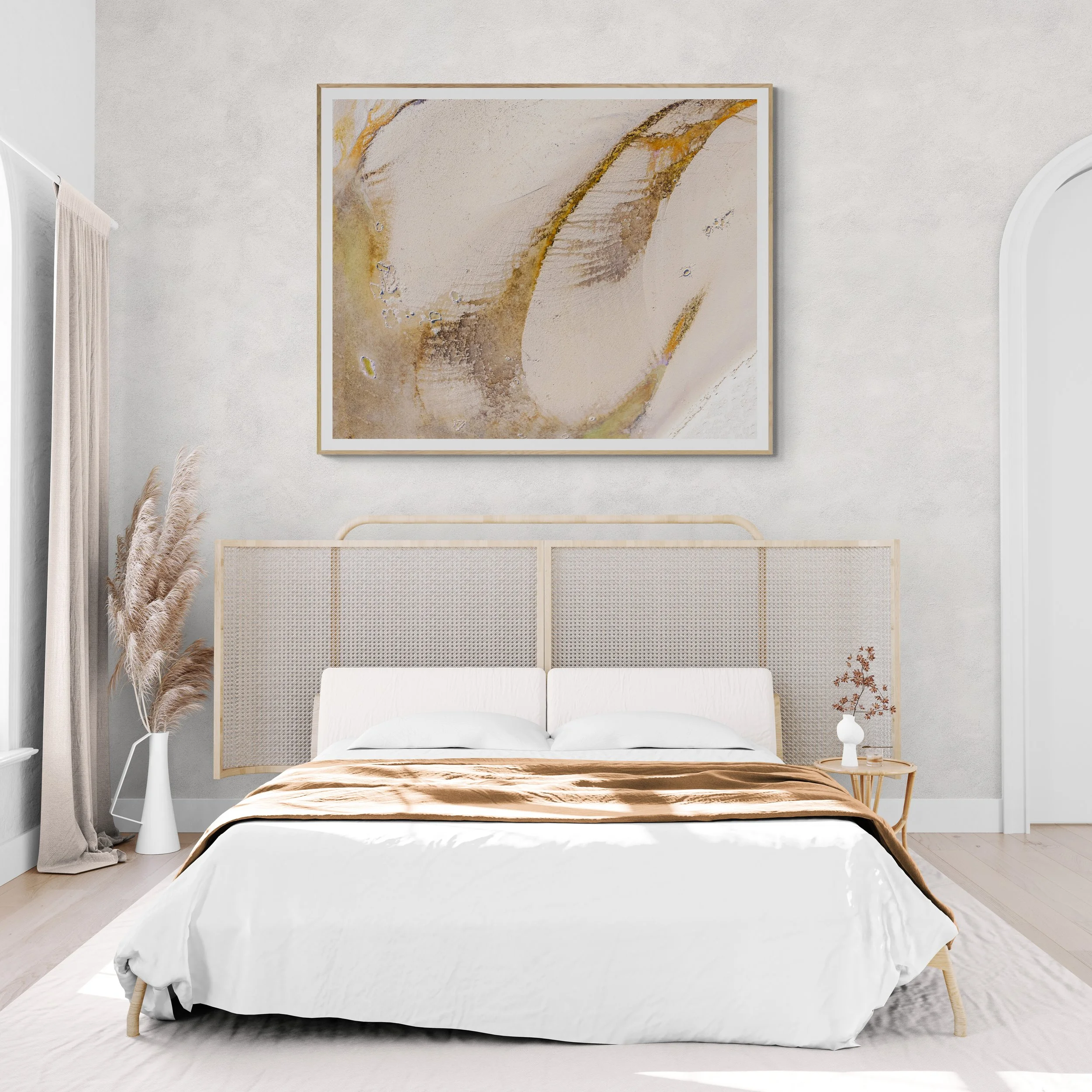 A minimalistic bedroom with a white bed, beige bedding, and a textured headboard, a vase with pampas grass on the left, a small side table with flowers on the right, a large abstract painting above the bed, and light-colored walls and flooring.