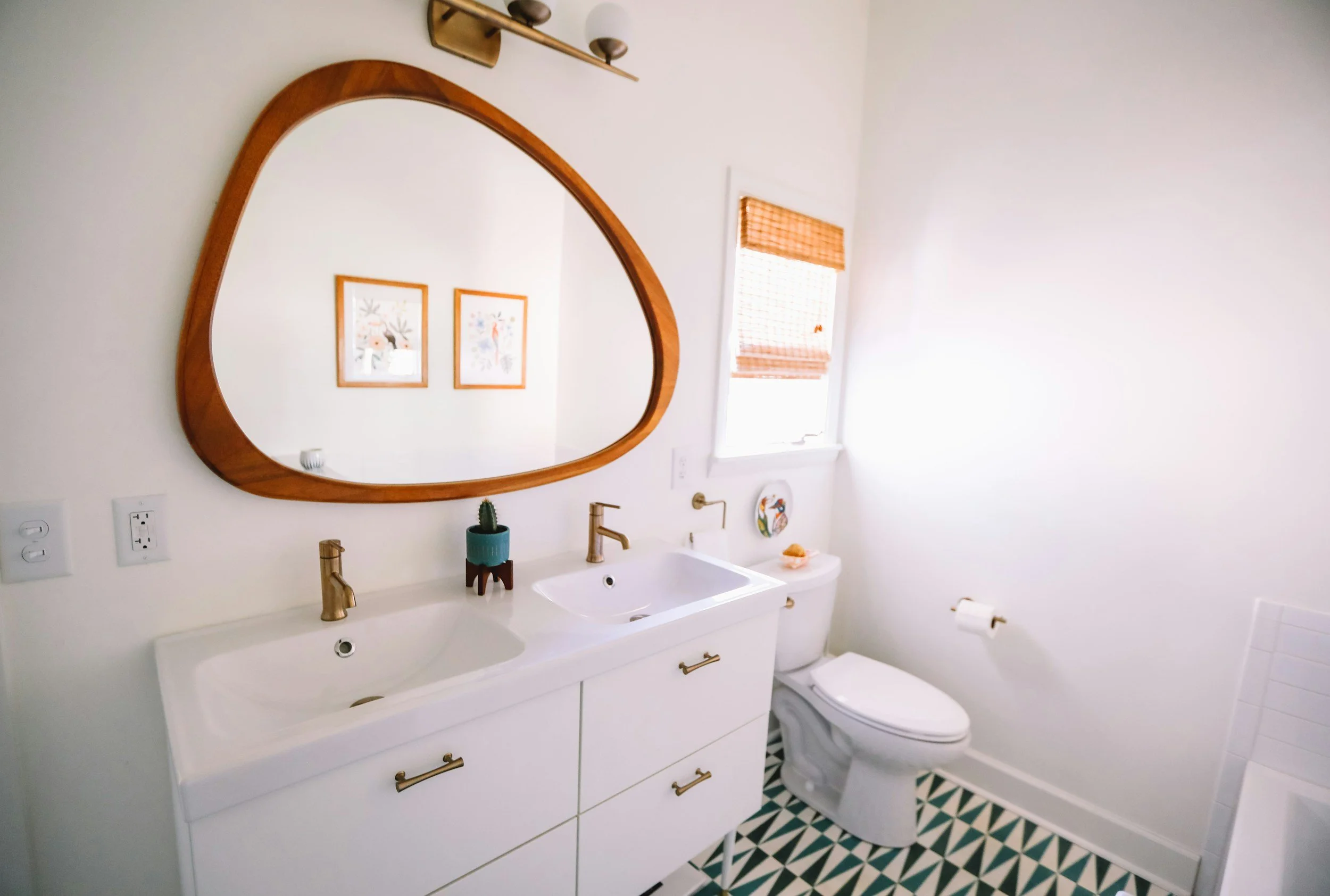 A modern bathroom with a white double sink vanity, brass fixtures, and a large, irregularly shaped wooden mirror. There is a cactus in a teal pot on the counter, above a white toilet. The wall has two framed art pieces and a small decorative dish. The floor features black, white, and beige geometric patterned tiles, and there is a small window with orange bamboo blinds.