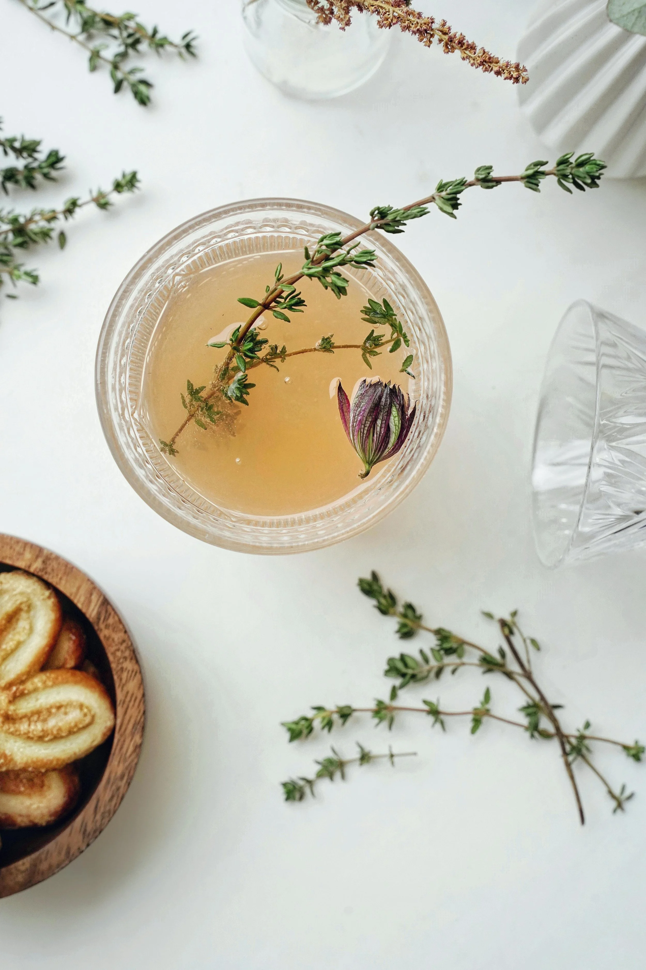 A glass of herbal tea with a sprig of thyme and a purple flower petal, surrounded by sprigs of thyme, with a wooden bowl of pinched cookies and empty glasses nearby on a white surface.