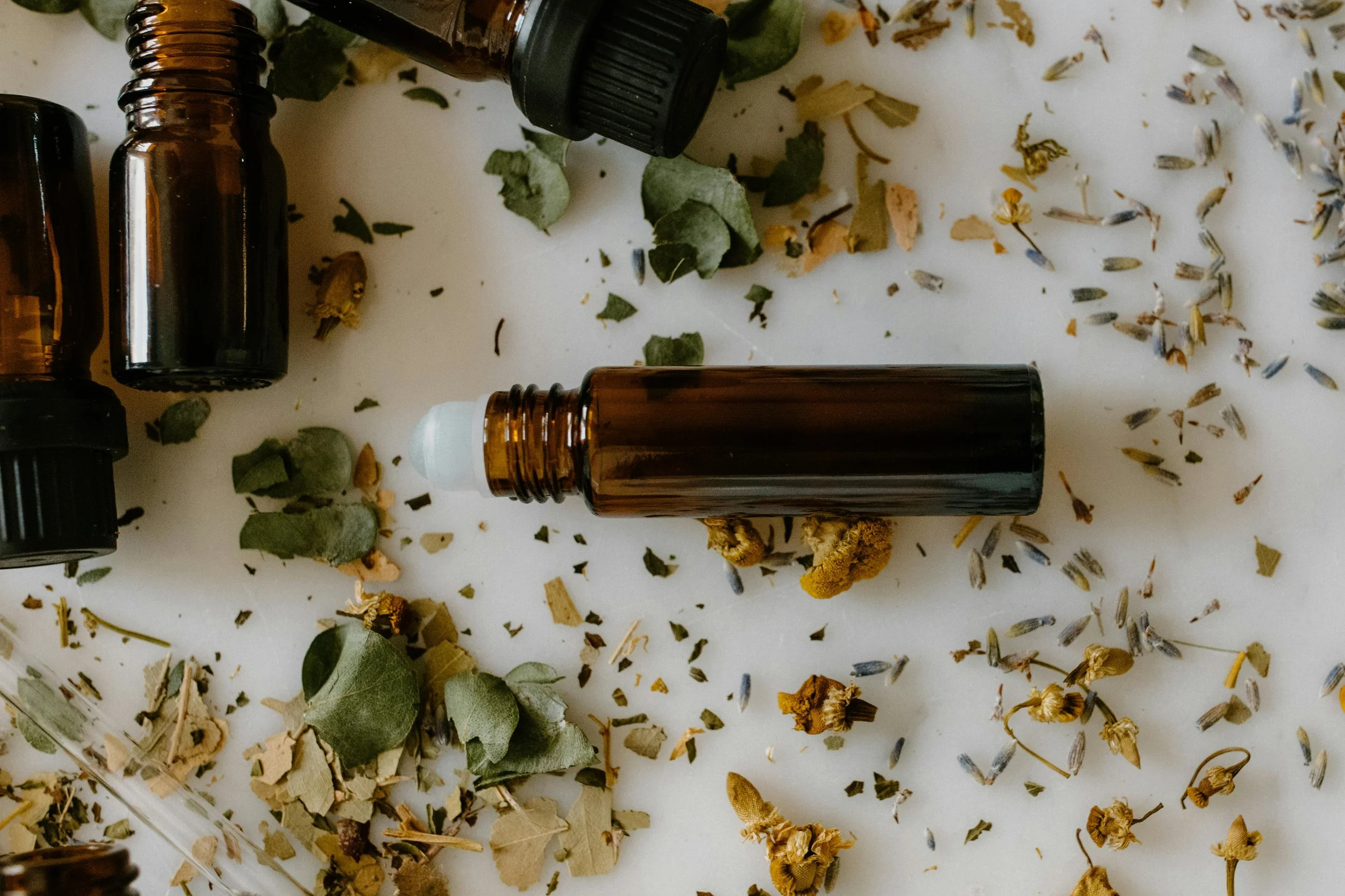 Amber glass essential oil bottles surrounded by dried herbs and flower petals on a white surface.