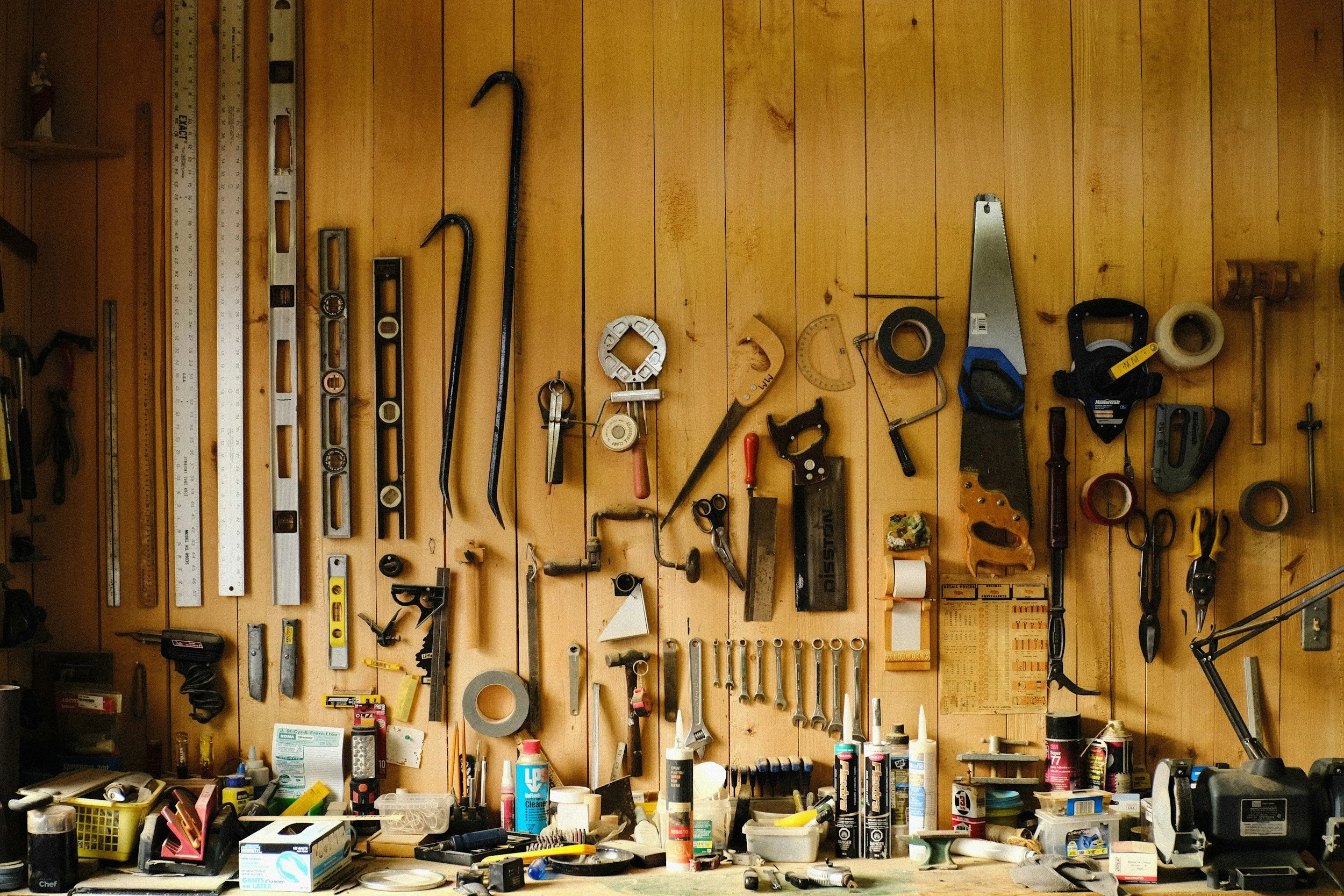 Wall with various hand tools hanging, including hammers, screwdrivers, wrenches, and measuring tools, with cluttered workbench below.