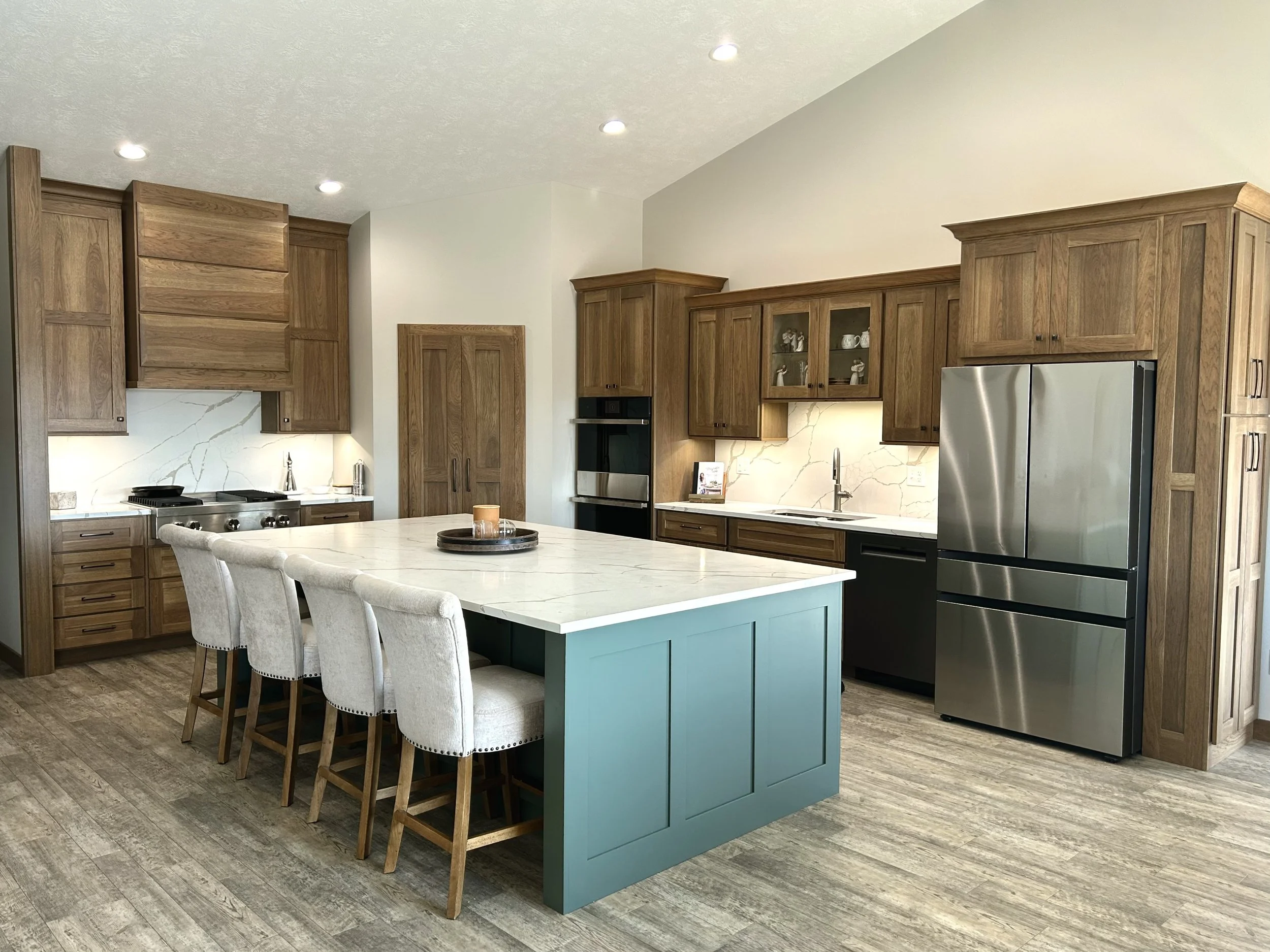 Modern kitchen with wooden cabinetry, a large island with a white marble countertop, beige upholstered chairs, stainless steel refrigerator, built-in oven, and a marble backsplash.