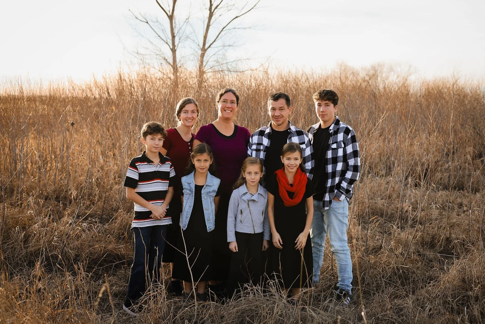 Family portrait of ten people, adults and children, standing in a field of brown grass during sunset.