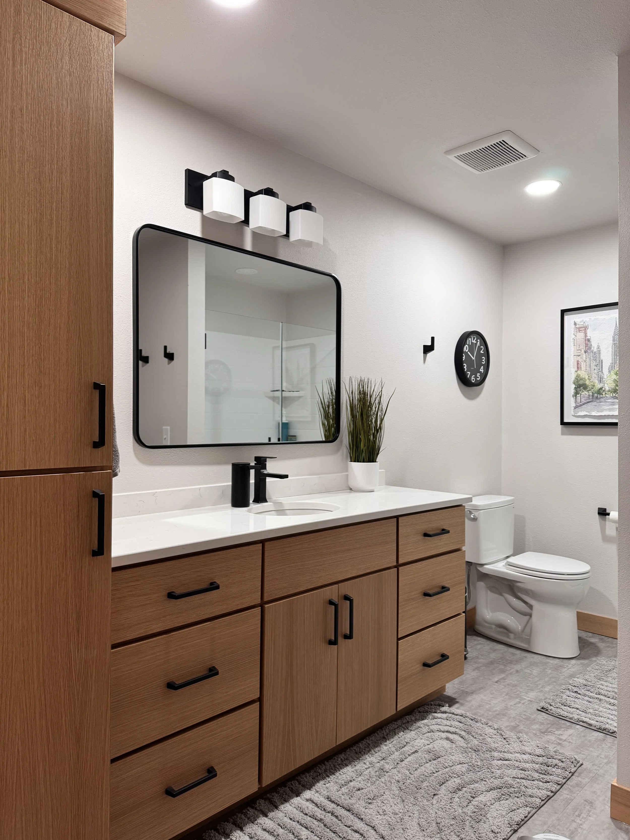 Modern bathroom with wooden vanity, white countertop, black fixtures, a mirror, a black wall clock, a plant, and a shower in the background.