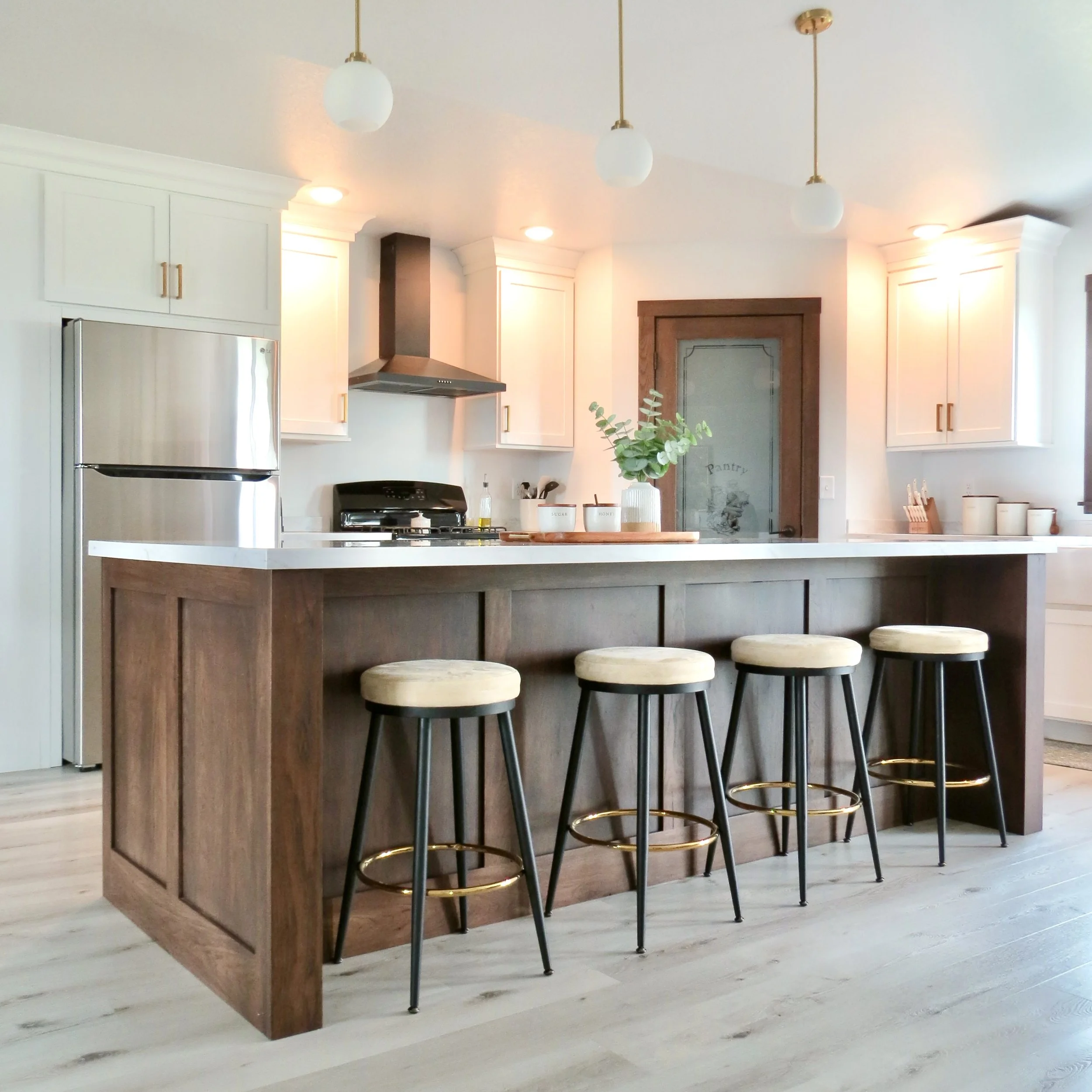Modern kitchen with white cabinets, stainless steel refrigerator, wooden kitchen island with four beige cushioned bar stools, pendant lights, and a chalkboard pantry door.