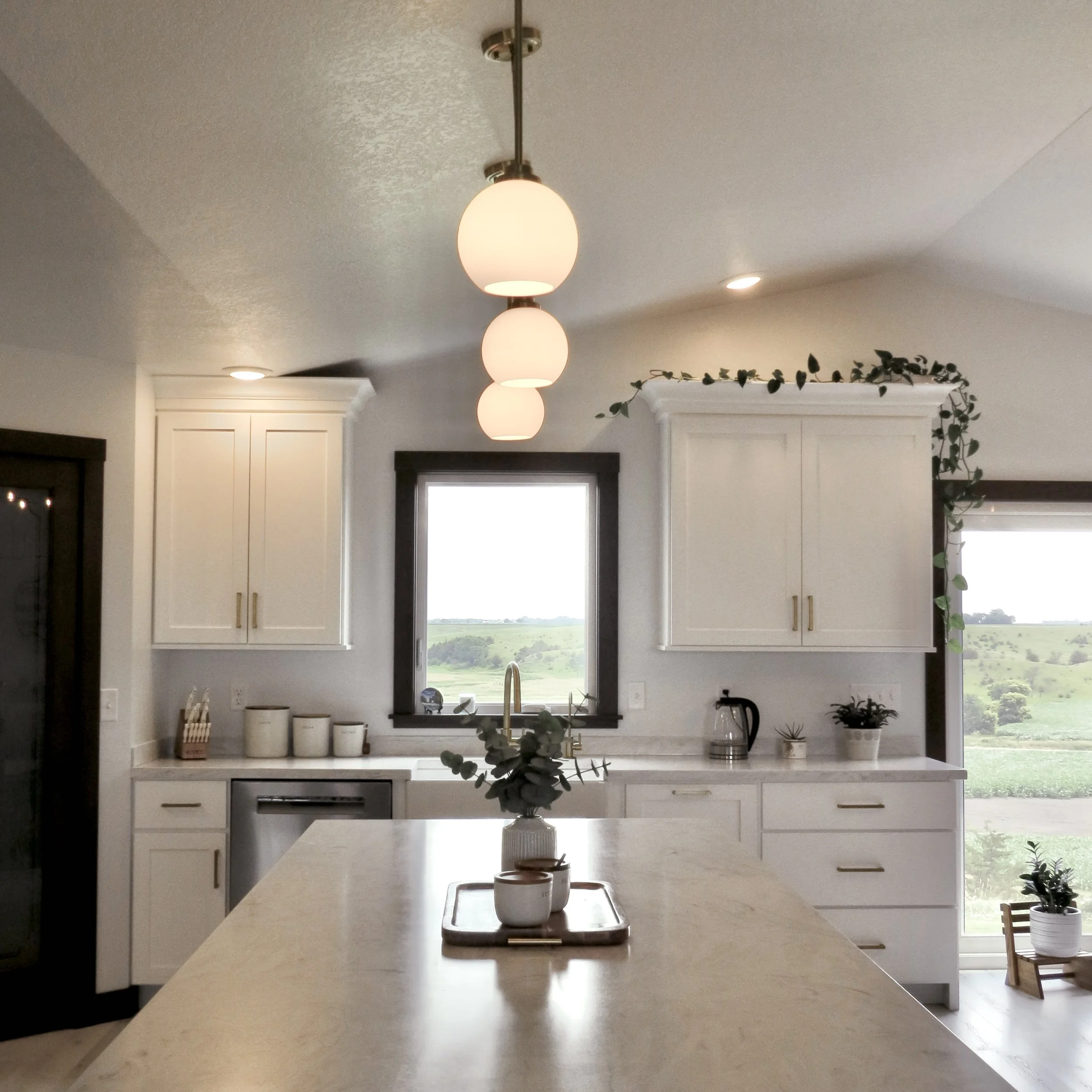 Modern kitchen with white cabinets, black window frames, pendant lights, and a view of green fields outside.