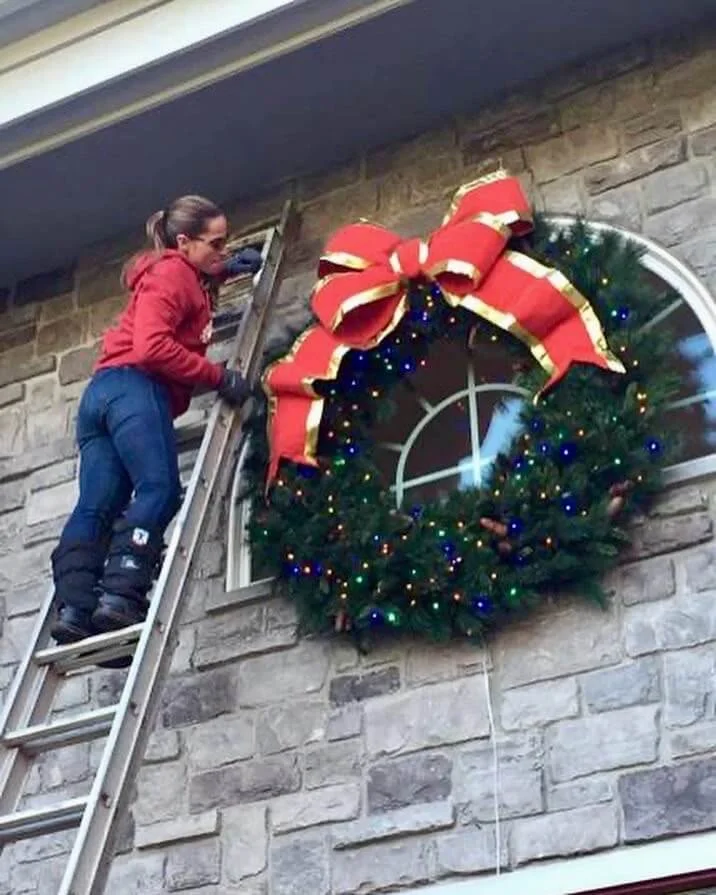 SHINE Owner Jenn installing a five-foot wreath with big red and gold bow and multicolor LED light strings on a home in Chittenango, NY in 2015