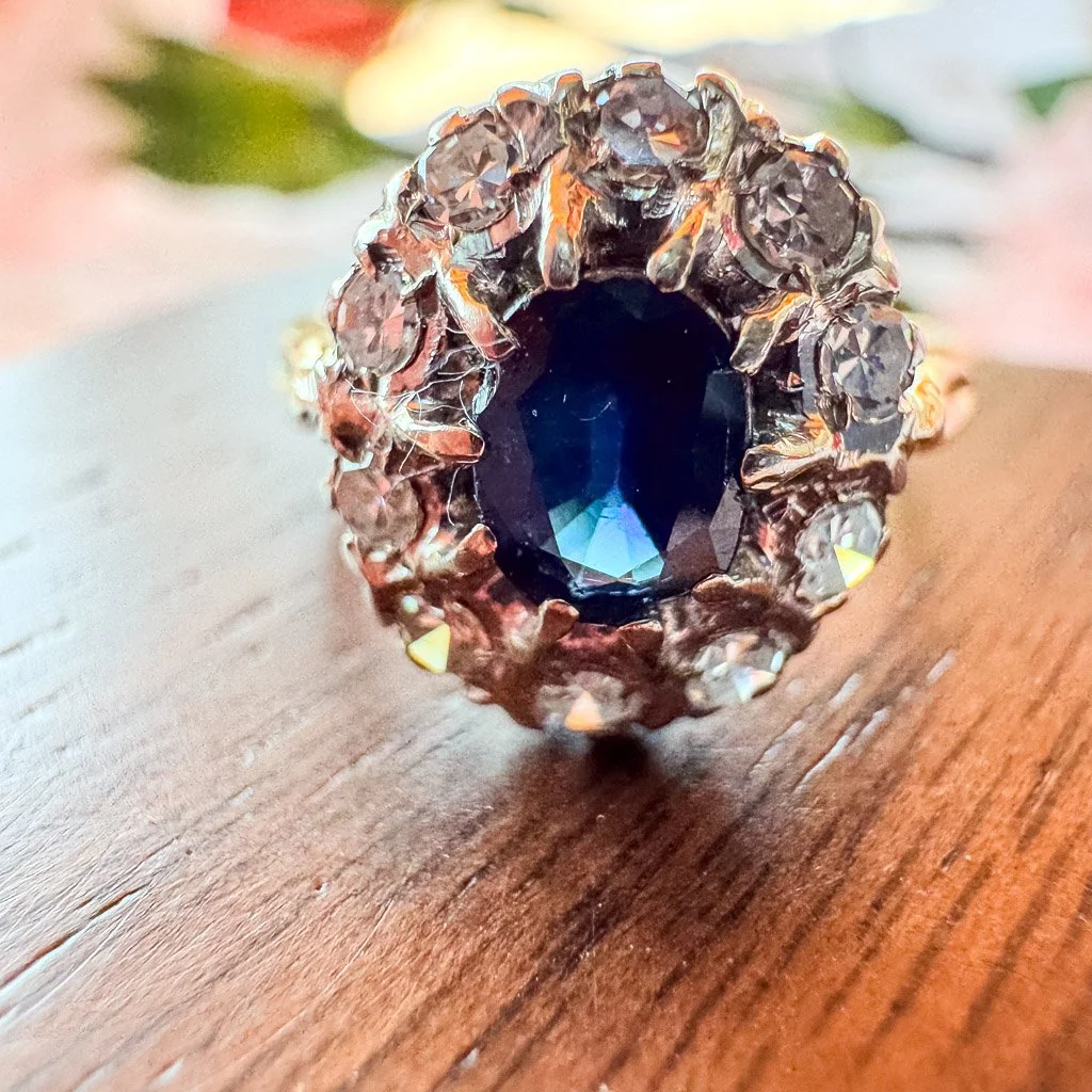 An Old CUt dark blue sapphire with old cut diamonds surrounding the sapphire in a halo. Ring has a gold band and is setting on a wood jewelry box. 