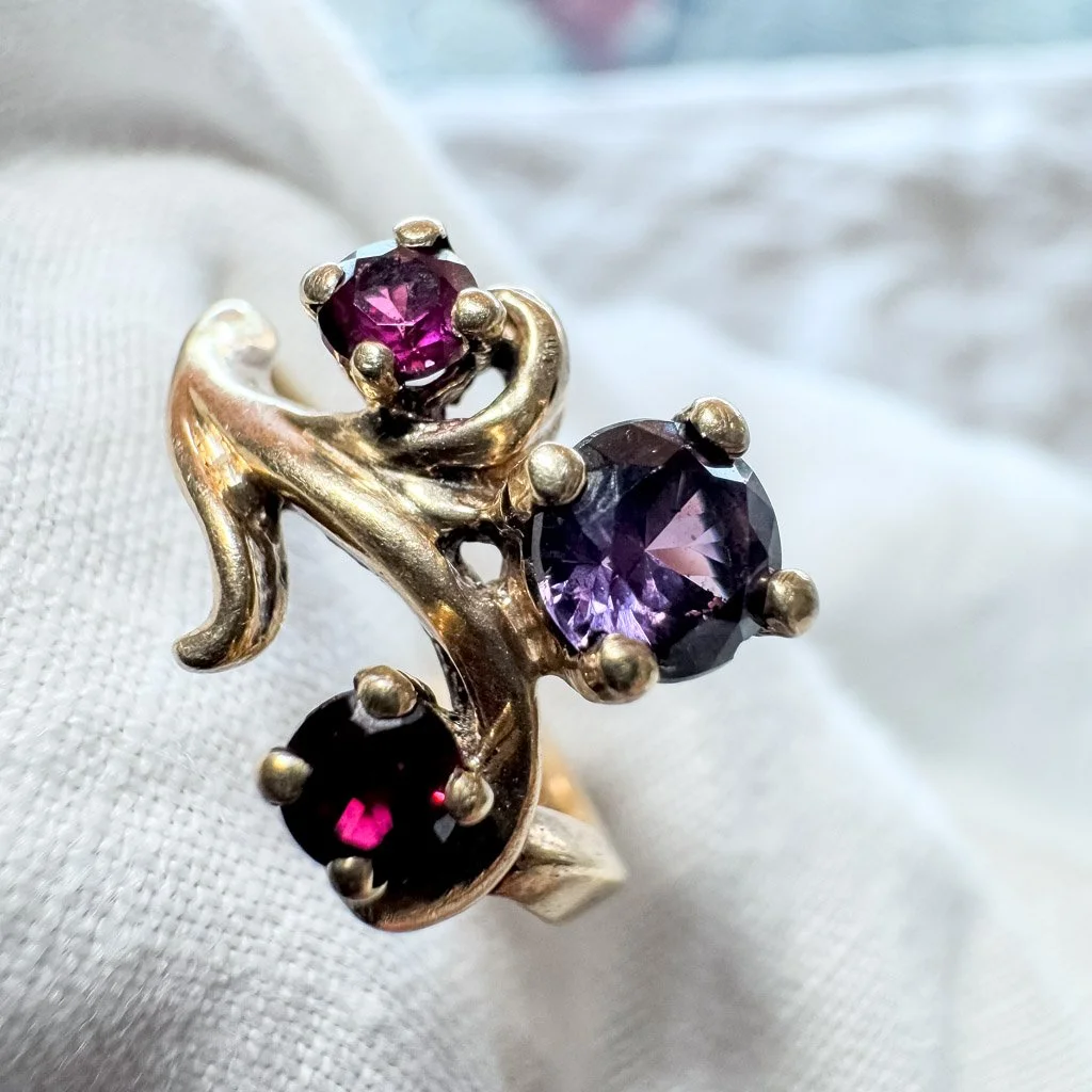 Gold ring with three purple and pink gemstones set in prongs, resting on white fabric.