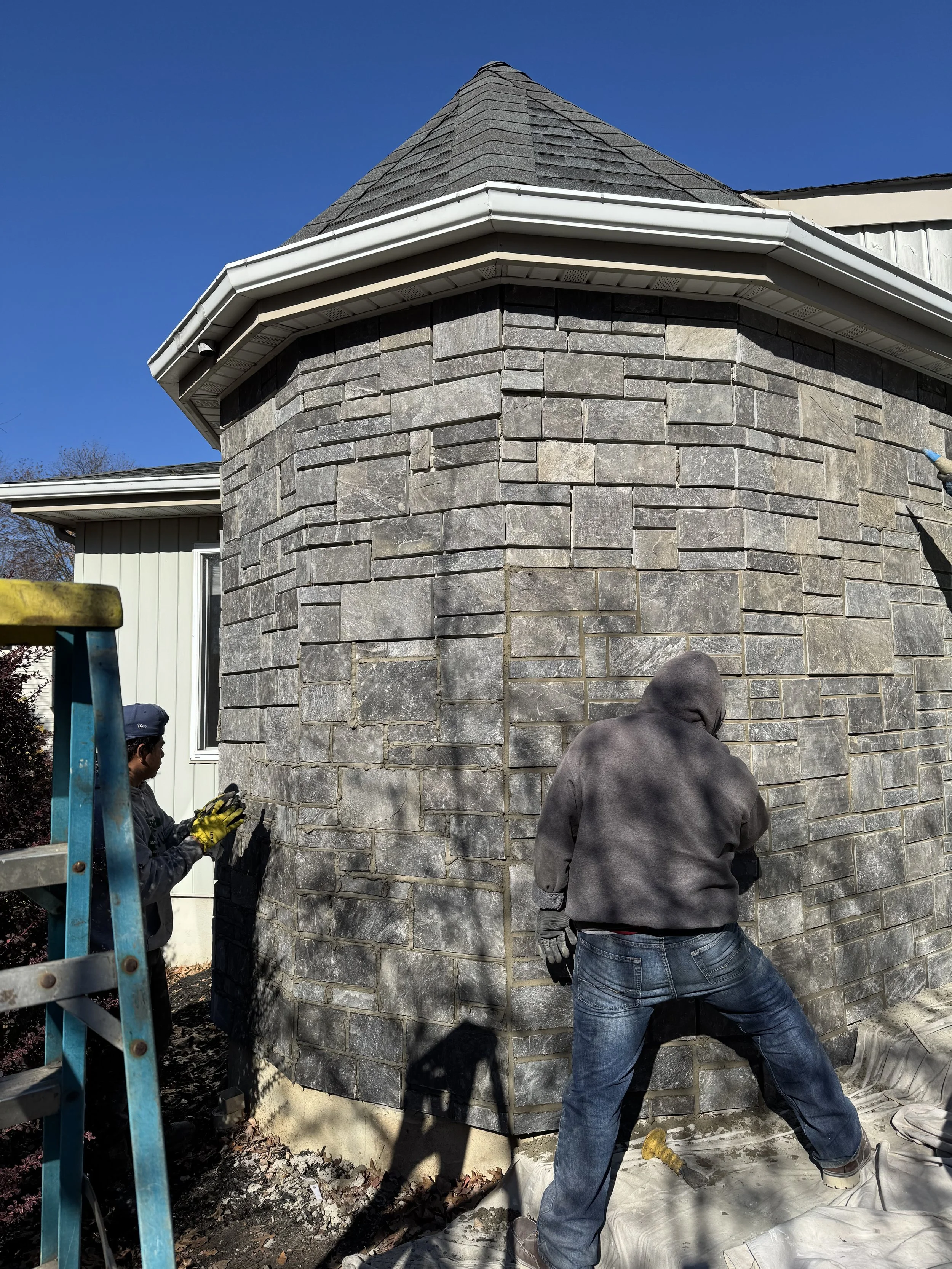 Two workers applying gray stone veneer to the exterior wall of a round turret on a house. One worker is standing with a hoodie and jeans, and the other is wearing gloves and a cap, working near a ladder.