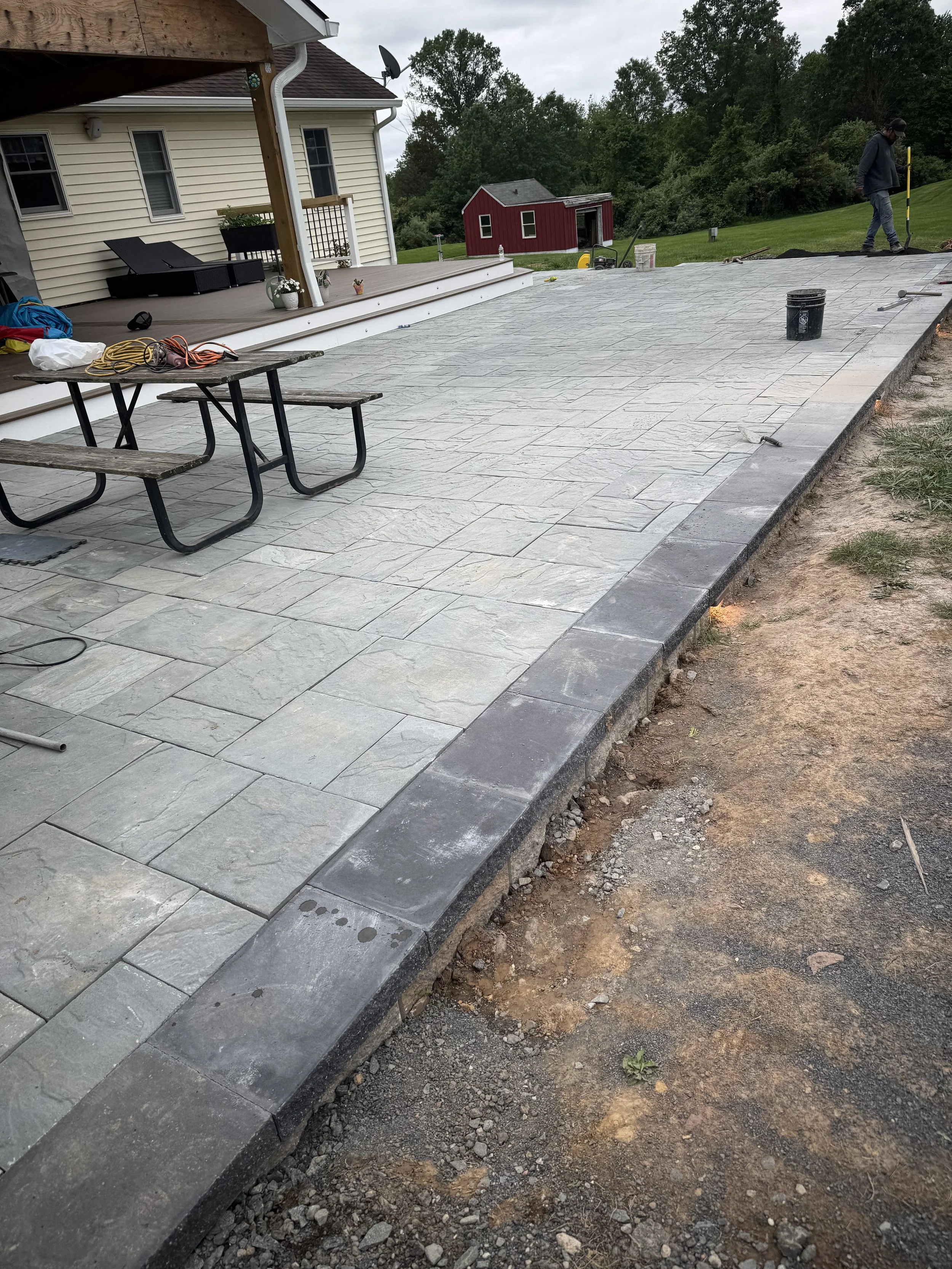 A newly constructed outdoor patio with stone pavers and a low brick border next to a house with a covered porch. The patio is in the process of being finished, with construction tools and materials visible. A person is working in the background, and there is a grassy yard with trees beyond.