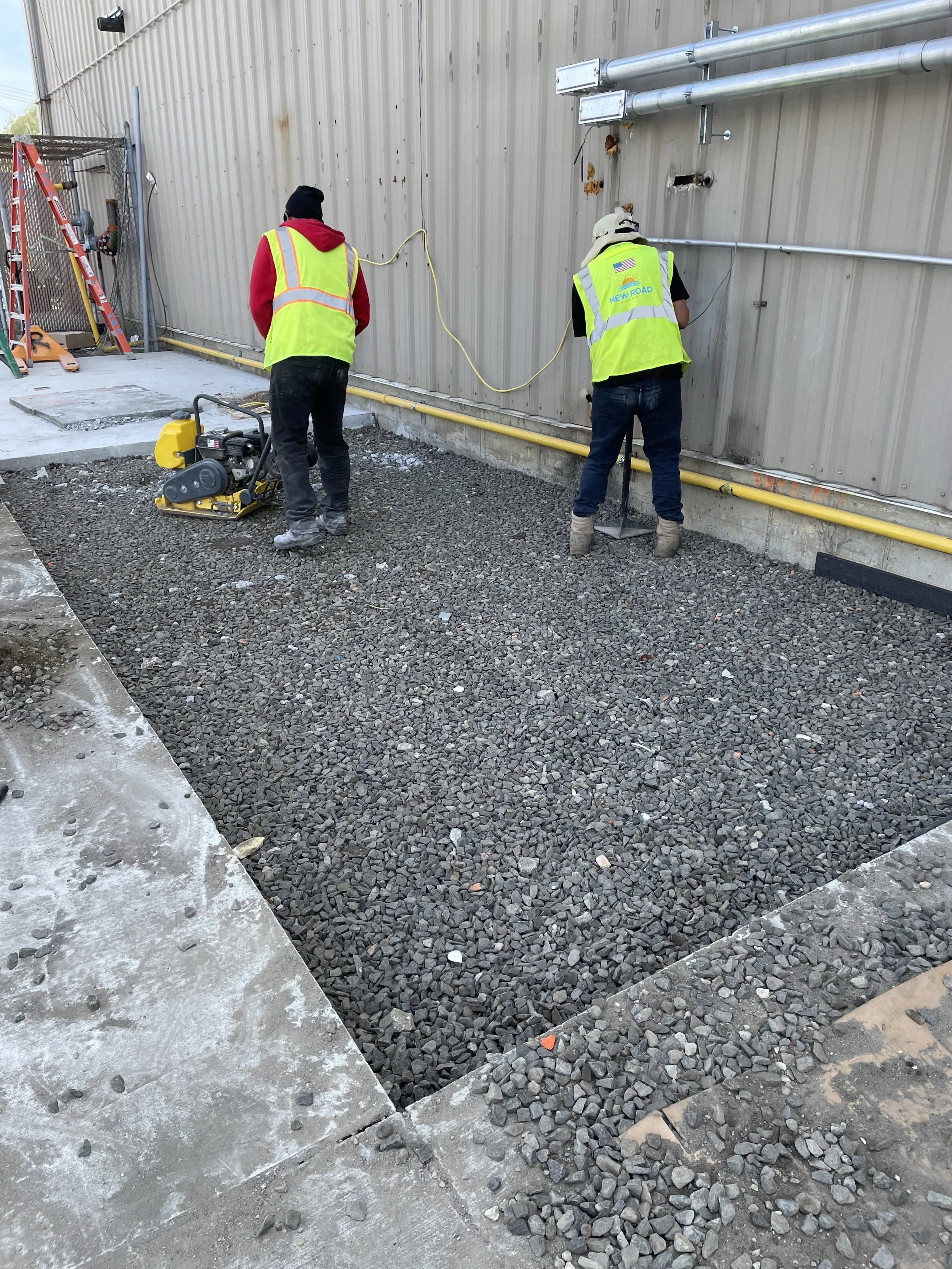 Two construction workers in safety vests using equipment to level gravel on a construction site perimeter.