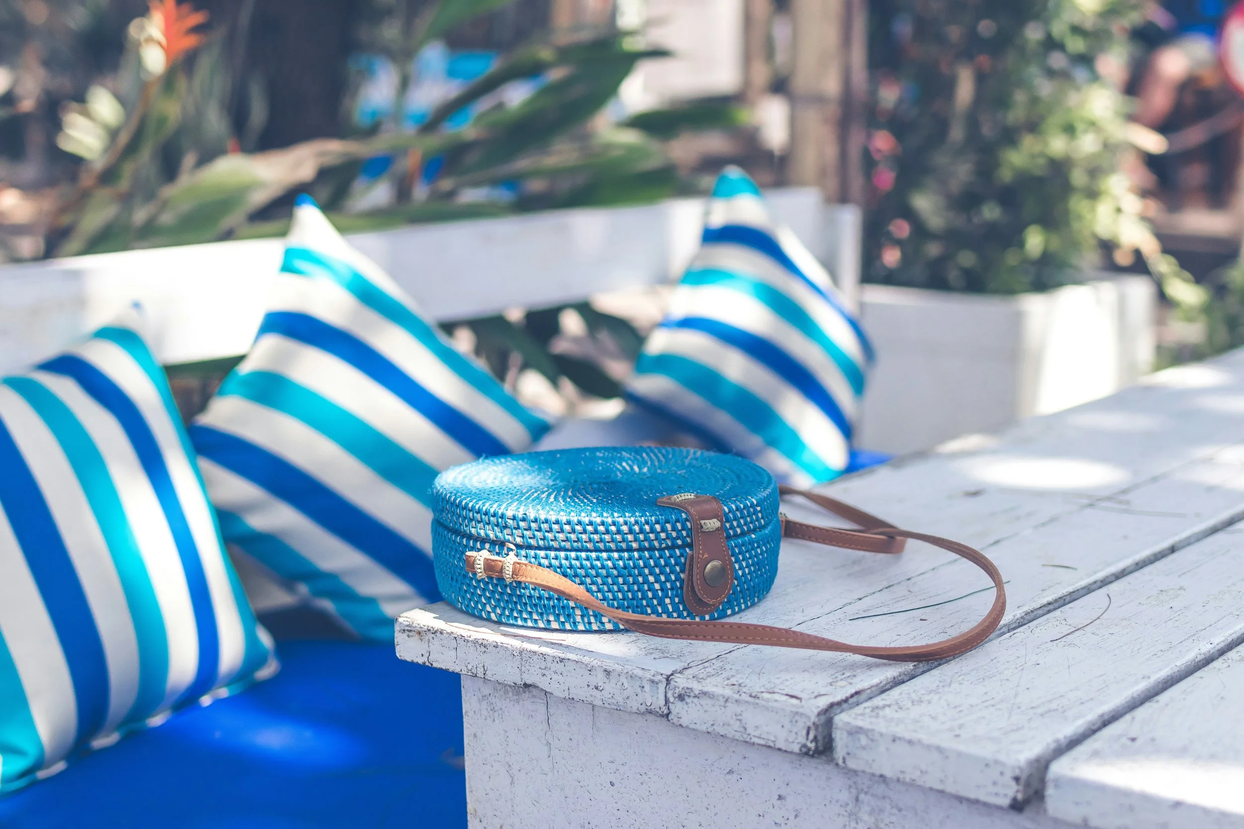Blue and white striped shoes on white wooden bench, with a blue woven handbag and sunglasses placed nearby, outdoors in a garden or patio.