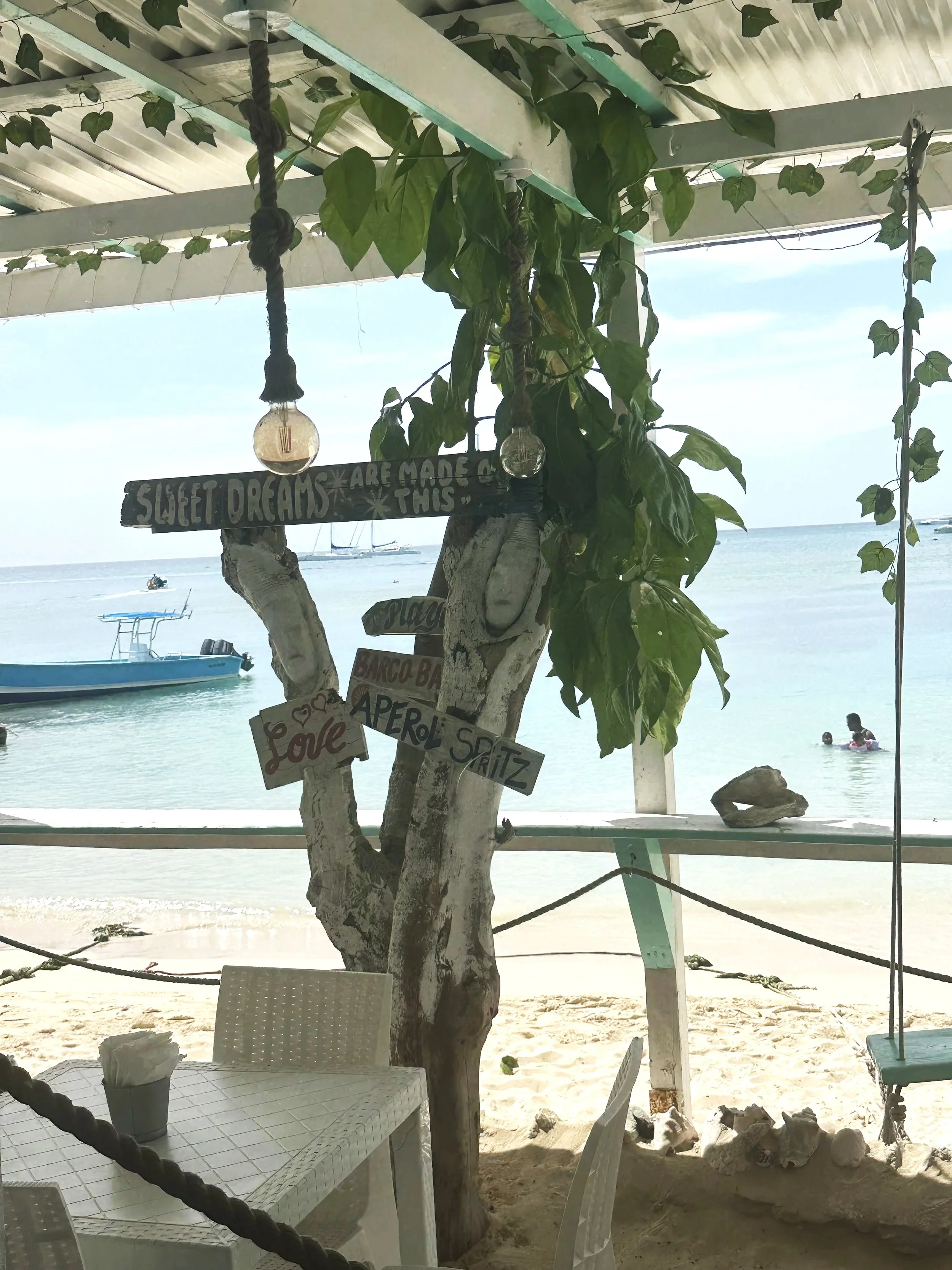 Beachside scene with a tree decorated with signs and hanging light bulbs. Several signs read 'Sweet Dreams Are Made Of This,' 'Love,' 'Playa,' 'Barco,' 'Aperol,' and 'Spritz.' In the background, the ocean is visible with boats and two people swimming, while indoors, a table with napkins is seen in the foreground.
