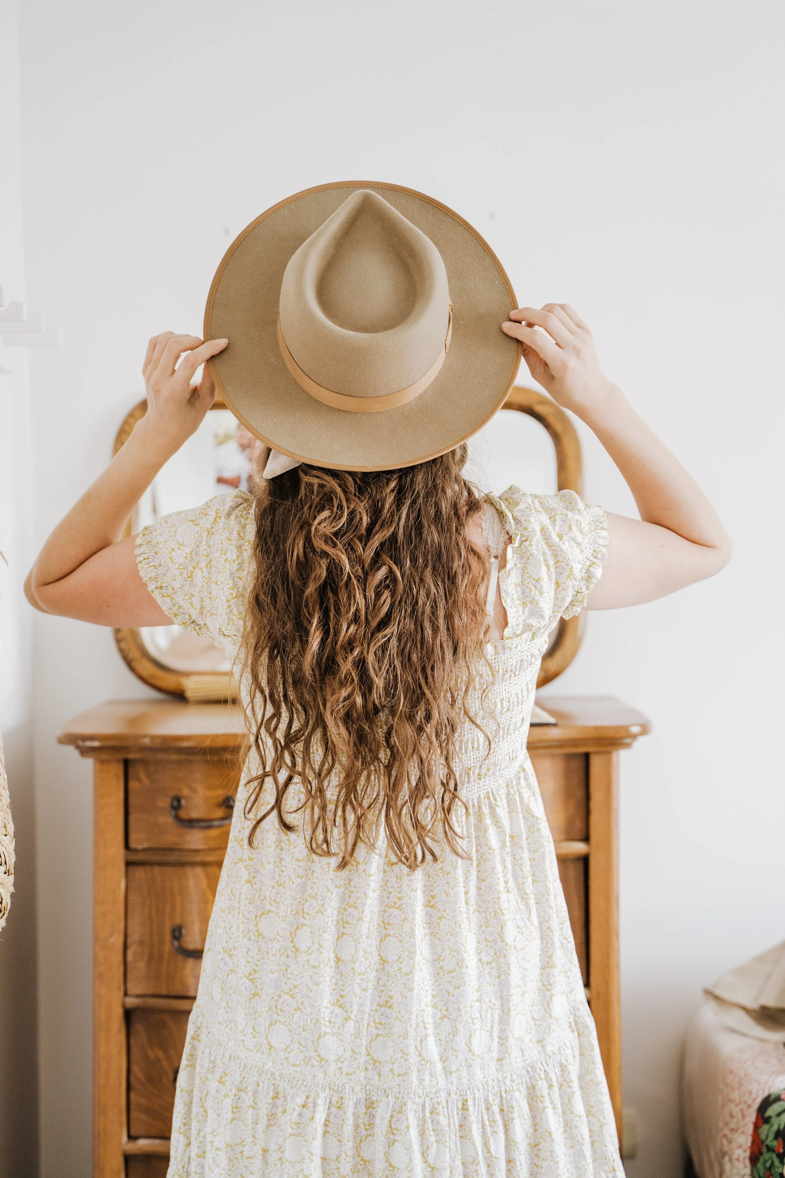 A woman with long, curly hair wears a light-colored, patterned dress and places a wide-brimmed, beige hat on her head. She is standing in a room with a wooden dresser against a white wall.