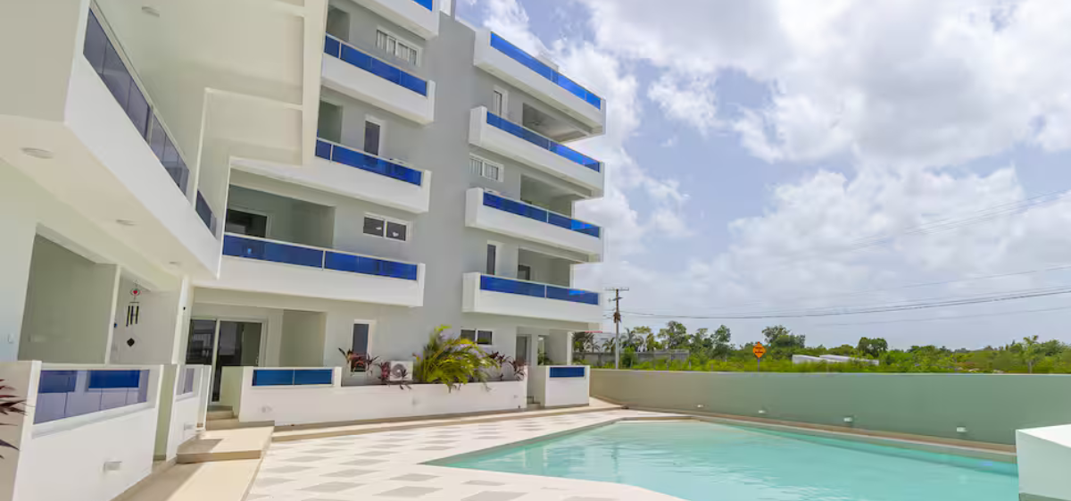 Modern multi-story apartment building with balconies and a swimming pool in the foreground, under partly cloudy sky.