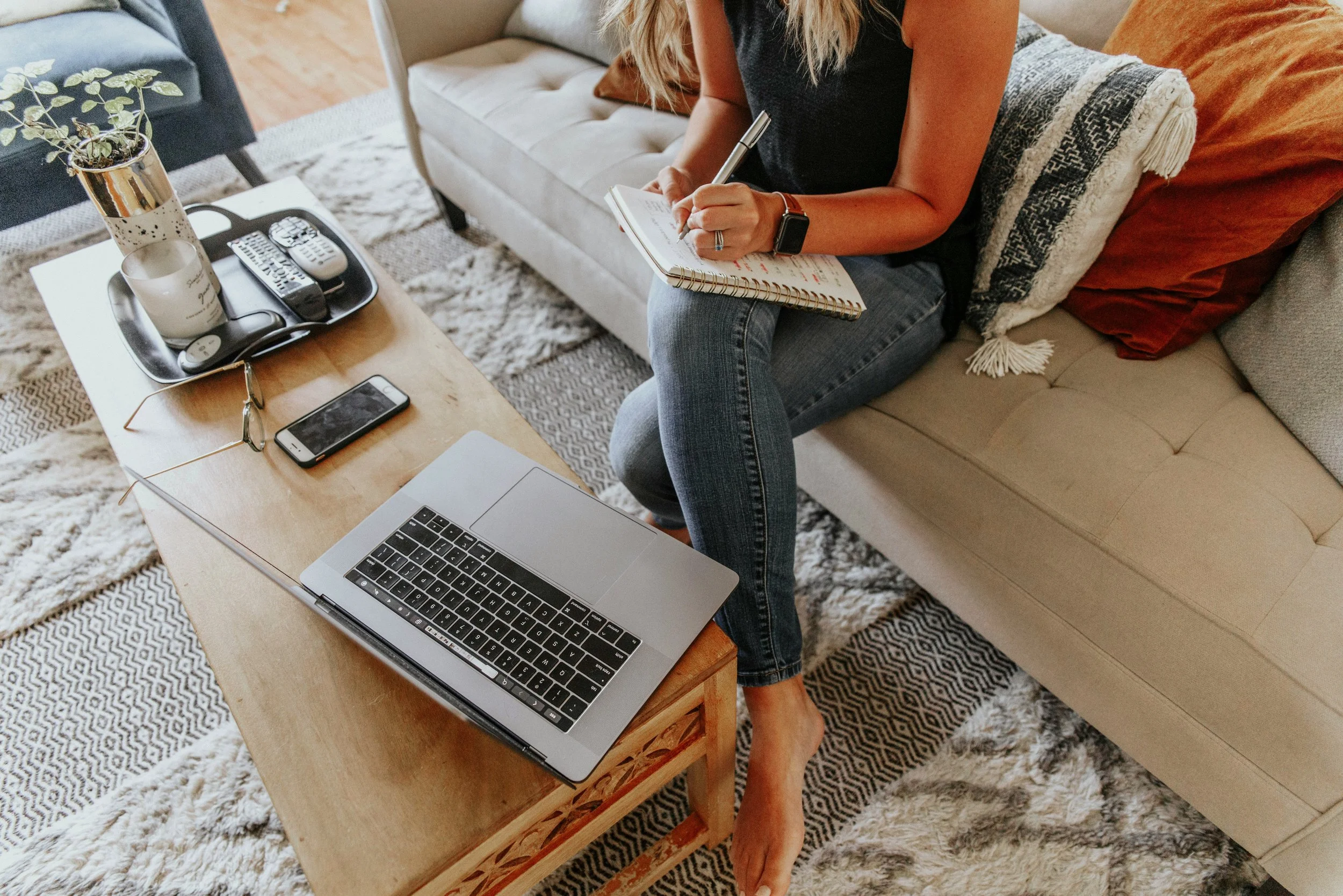 A woman sitting on a beige couch with colorful pillows, writing in a notebook with a pen. In front of her is a wooden coffee table with a silver laptop, a smartphone, sunglasses, and a tray with a plant, a candle, and remote controls. The setting appears cozy and casual in a home environment.