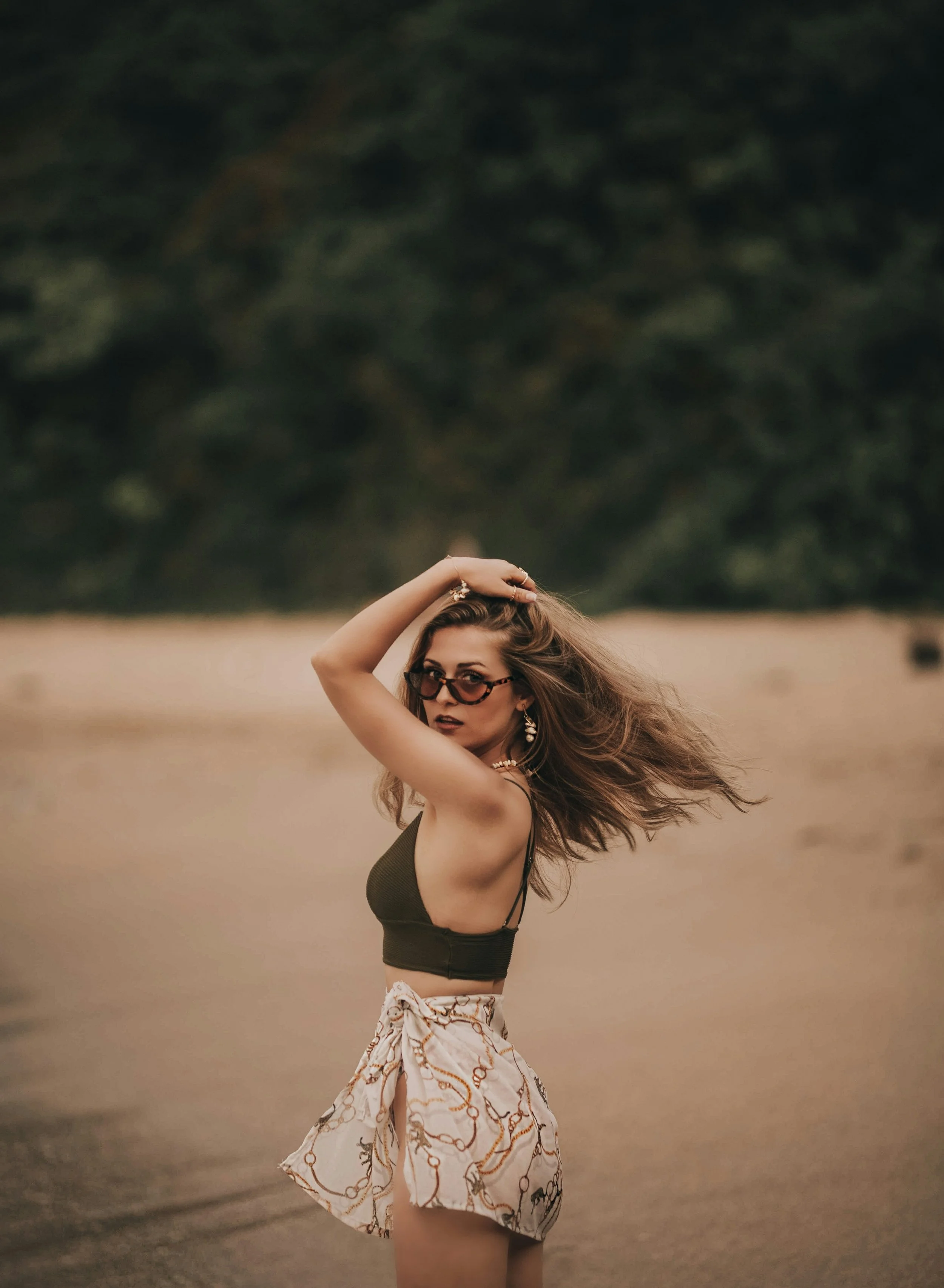 A woman standing in a field with trees in the background, wearing sunglasses, a green crop top, and a patterned skirt.