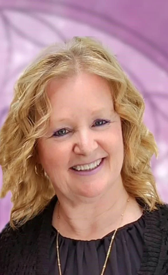 A smiling woman with wavy blonde hair wearing a black top and a gold necklace, standing against a pink and purple abstract background.