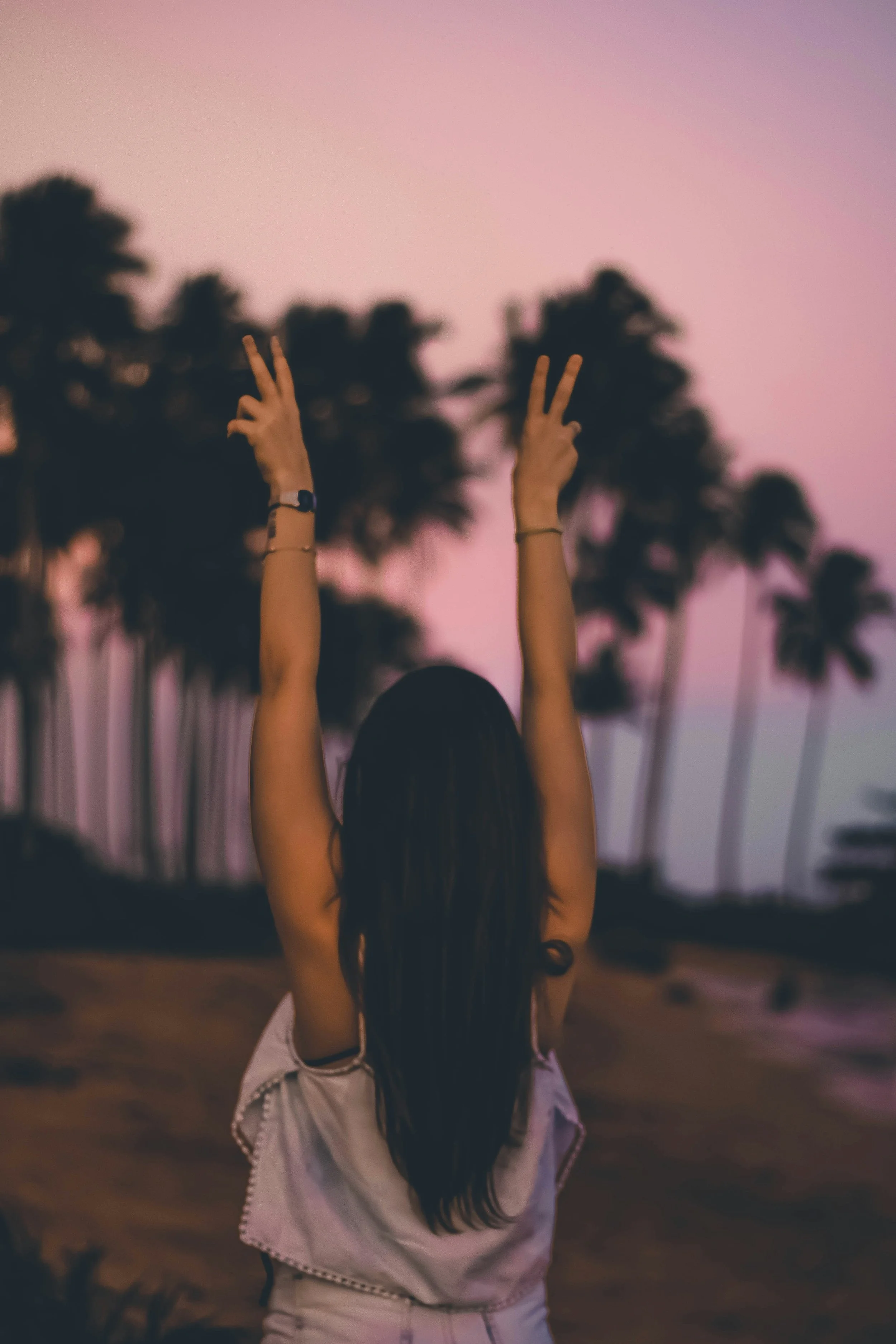 A girl with long dark hair is standing outdoors at sunset, raising her hands in the shape of peace signs against a backdrop of pink and purple sky with tall palm trees.