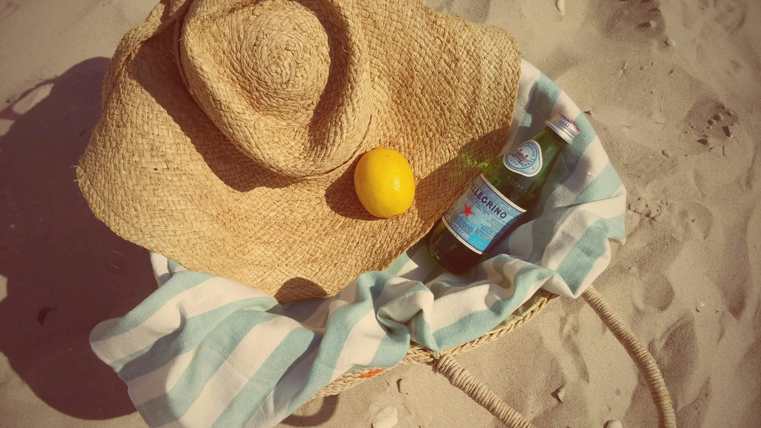 A straw hat, a lemon, a bottle of San Pellegrino sparkling water, and a light-colored cloth on a chair on the sand at the beach.