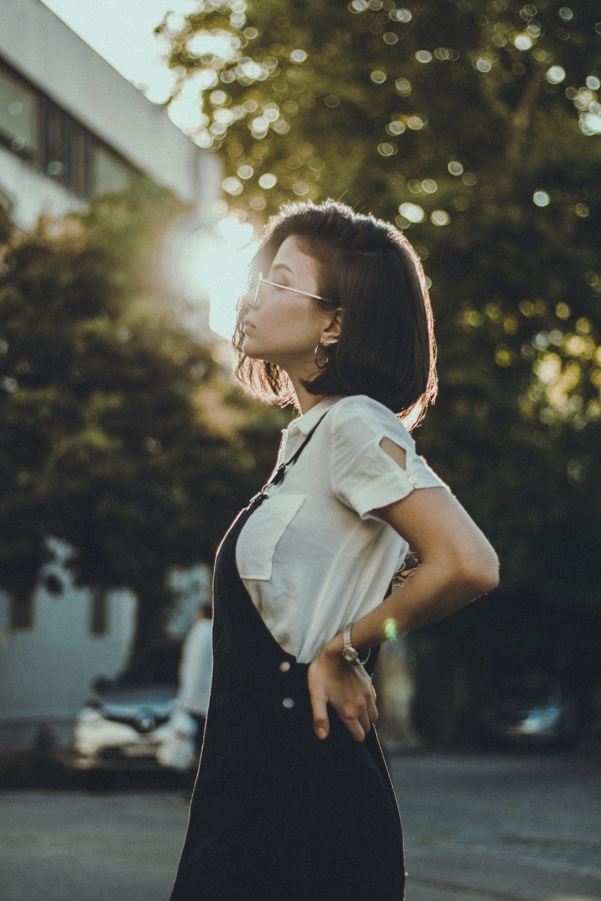 A woman with short dark hair, wearing sunglasses and a white shirt with cut-out sleeves, stands outdoors in the evening sunlight with trees and buildings in the background.