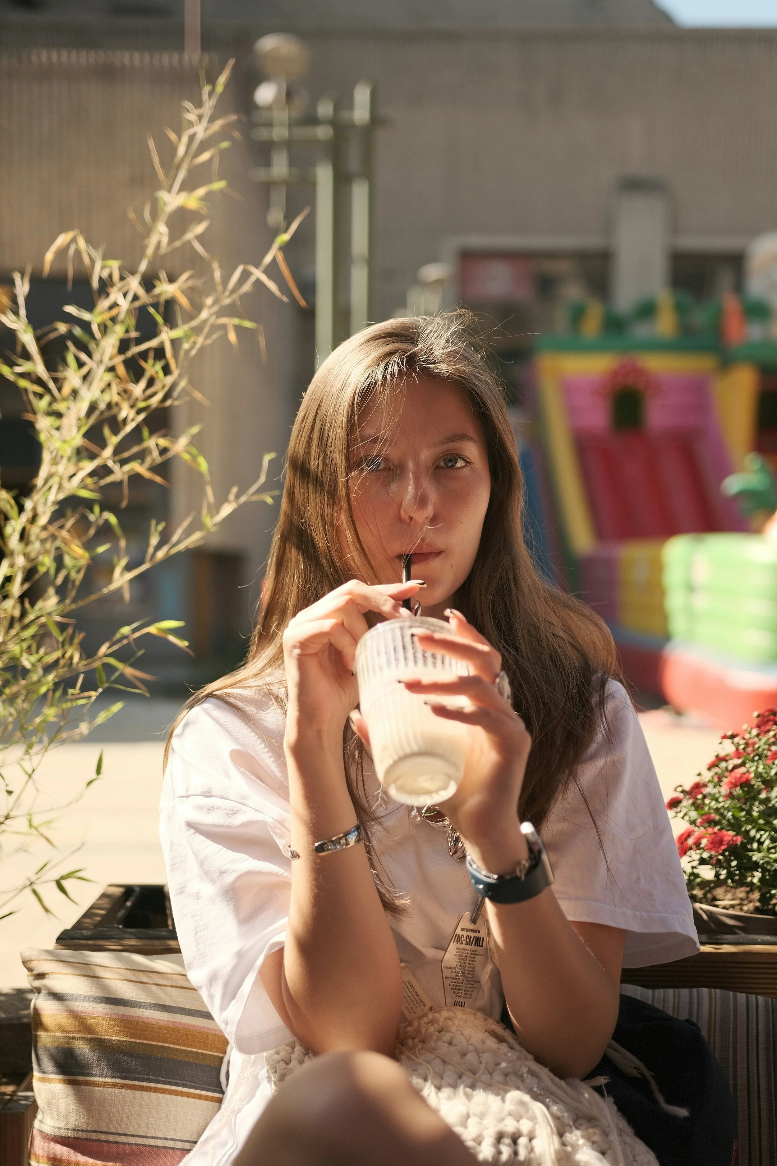 A young woman with long brown hair is sitting outdoors, drinking a beverage with a straw from a clear plastic cup. The scene is sunny with a blurred colorful amusement park ride in the background and some greenery to the side.