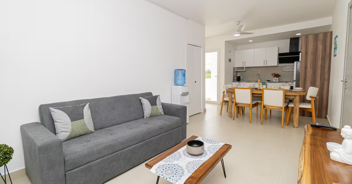 Living room with a gray sofa, a small wooden coffee table with a decorative cloth and bowl, and a sideboard with decorative items. Open kitchen with white cabinets, a dining table with six chairs, and a water dispenser.