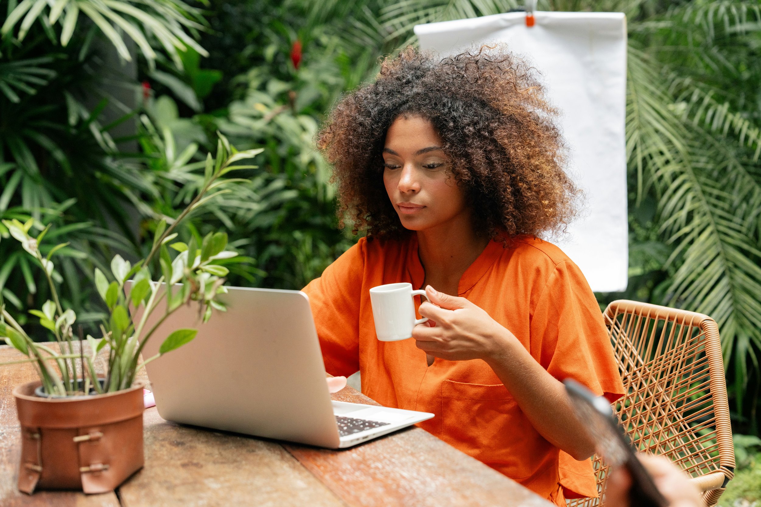A woman with curly hair wearing an orange top, sitting at a table in a lush green indoor garden, holding a white coffee mug, with a laptop in front of her.