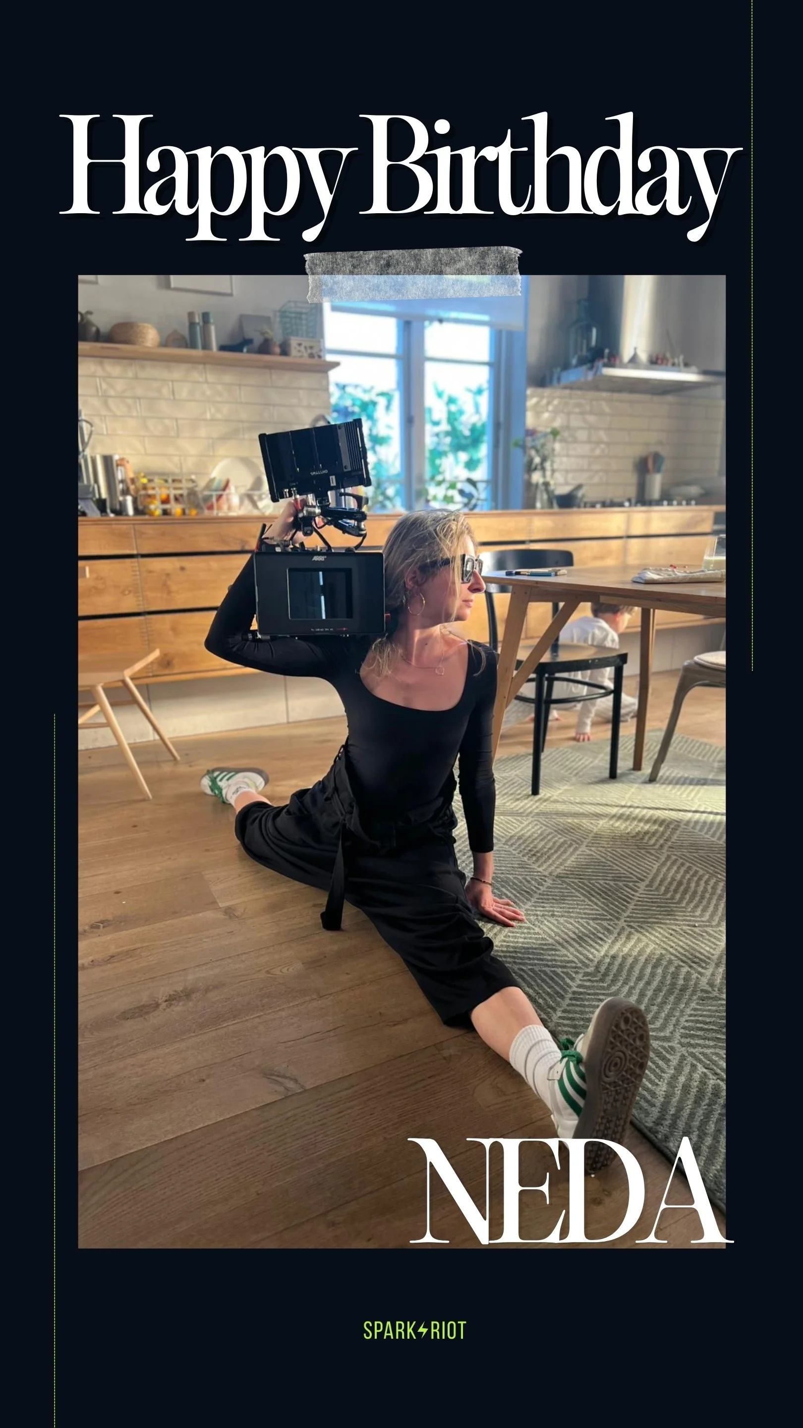 A woman in black clothing and sunglasses is performing a split on a wooden floor in a kitchen, holding a camera with a large lens on her shoulder. Text on the image says "Happy Birthday Neda" with a logo "SPARK RIOT" at the bottom.