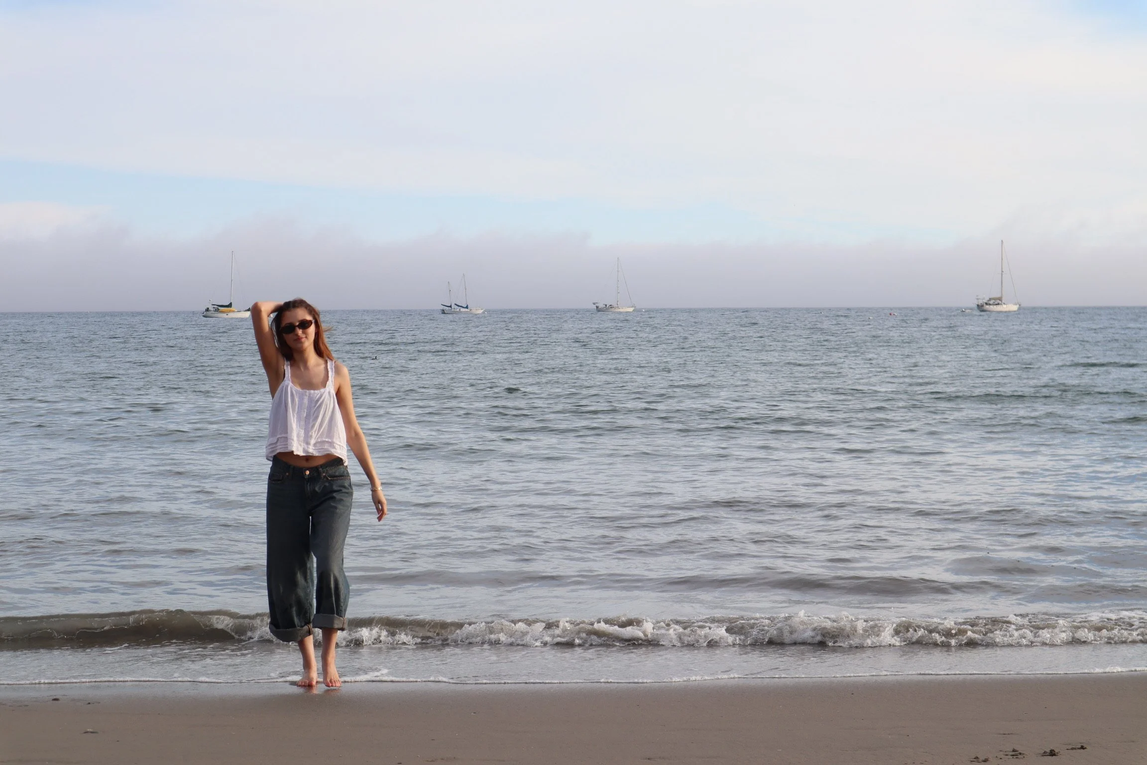 A woman with long hair wearing sunglasses, a white sleeveless top, and rolled-up jeans walking barefoot on the sandy beach with water and sailboats in the background.
