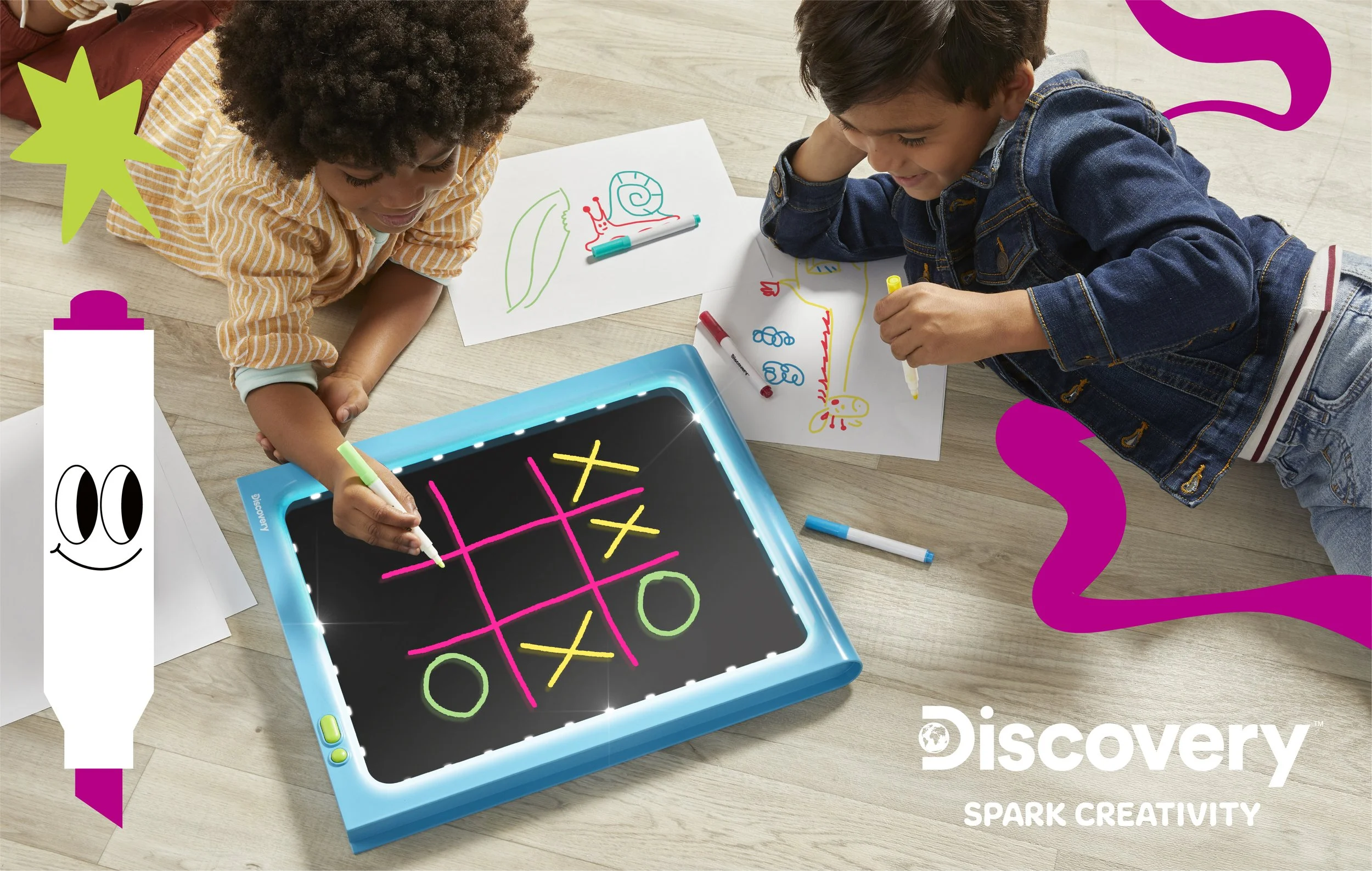 Two children playing a game of tic-tac-toe on an electronic tablet, surrounded by drawings and colorful illustrations, on a wooden floor.