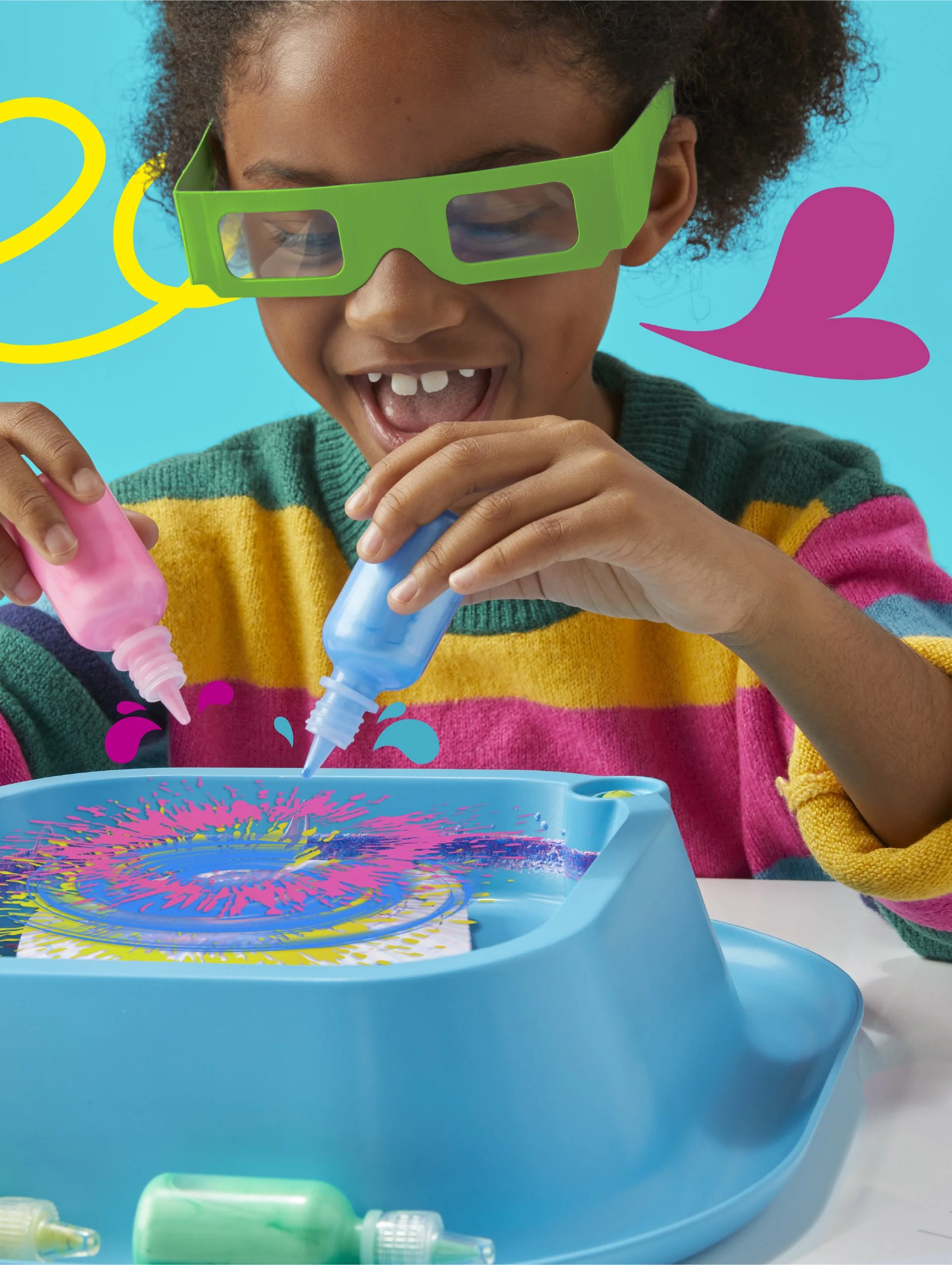 A smiling young girl wearing green glasses is holding two syringes, one pink and one blue, with colorful paint splashes on a tray in front of her.