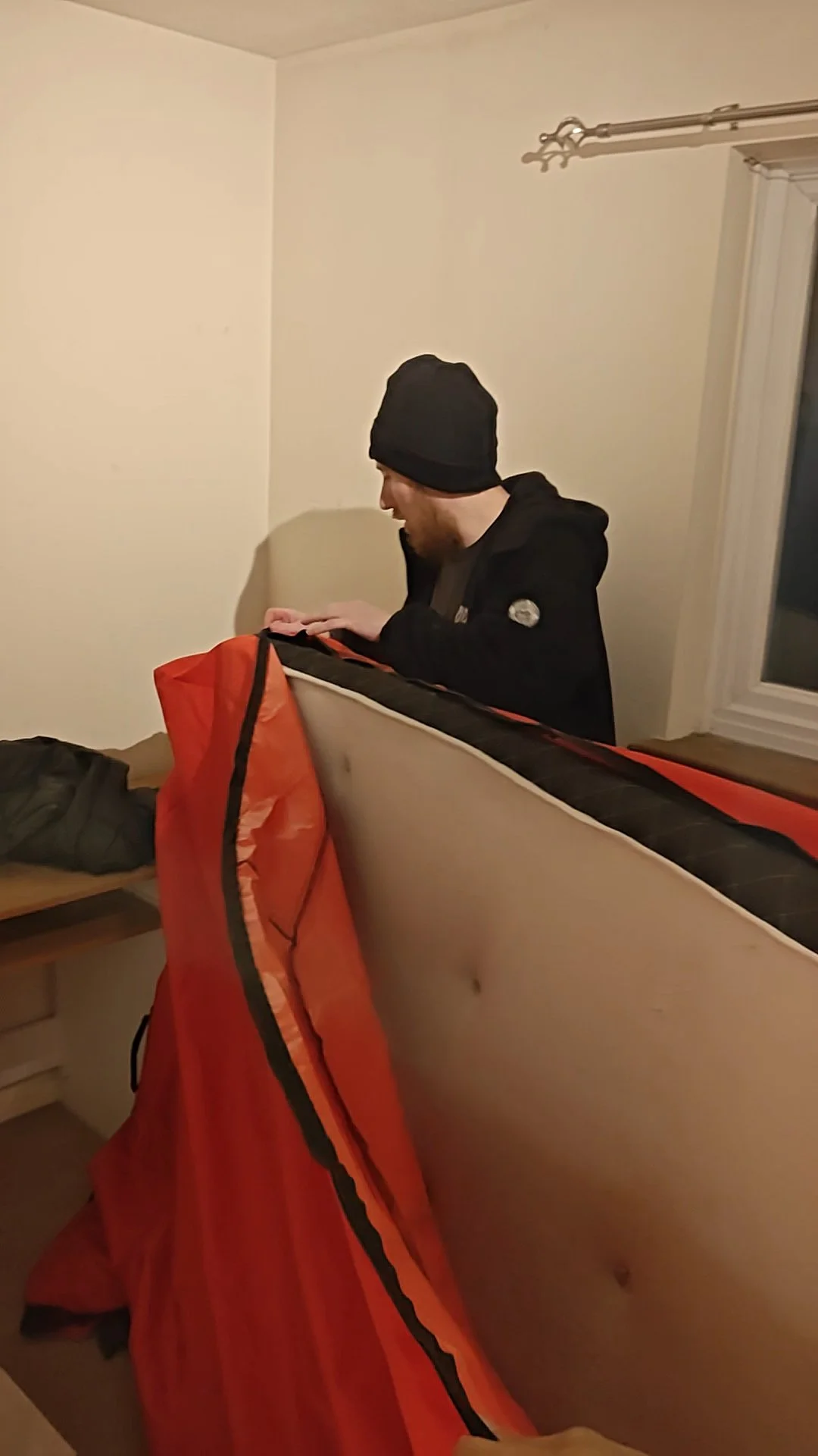 A man wearing a black beanie and black jacket standing indoors, looking down at an orange sleeping bag draped over a piece of furniture, with a window in the background.